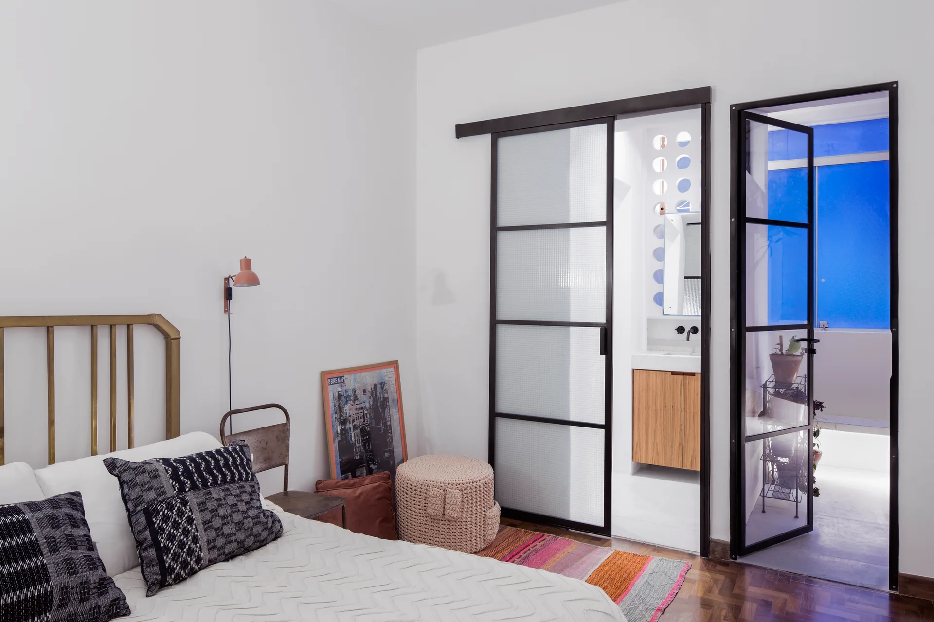 A bedroom featuring a brass bed, a pink wall sconce, and a black metal sliding glass door leading to an adjacent bathroom.