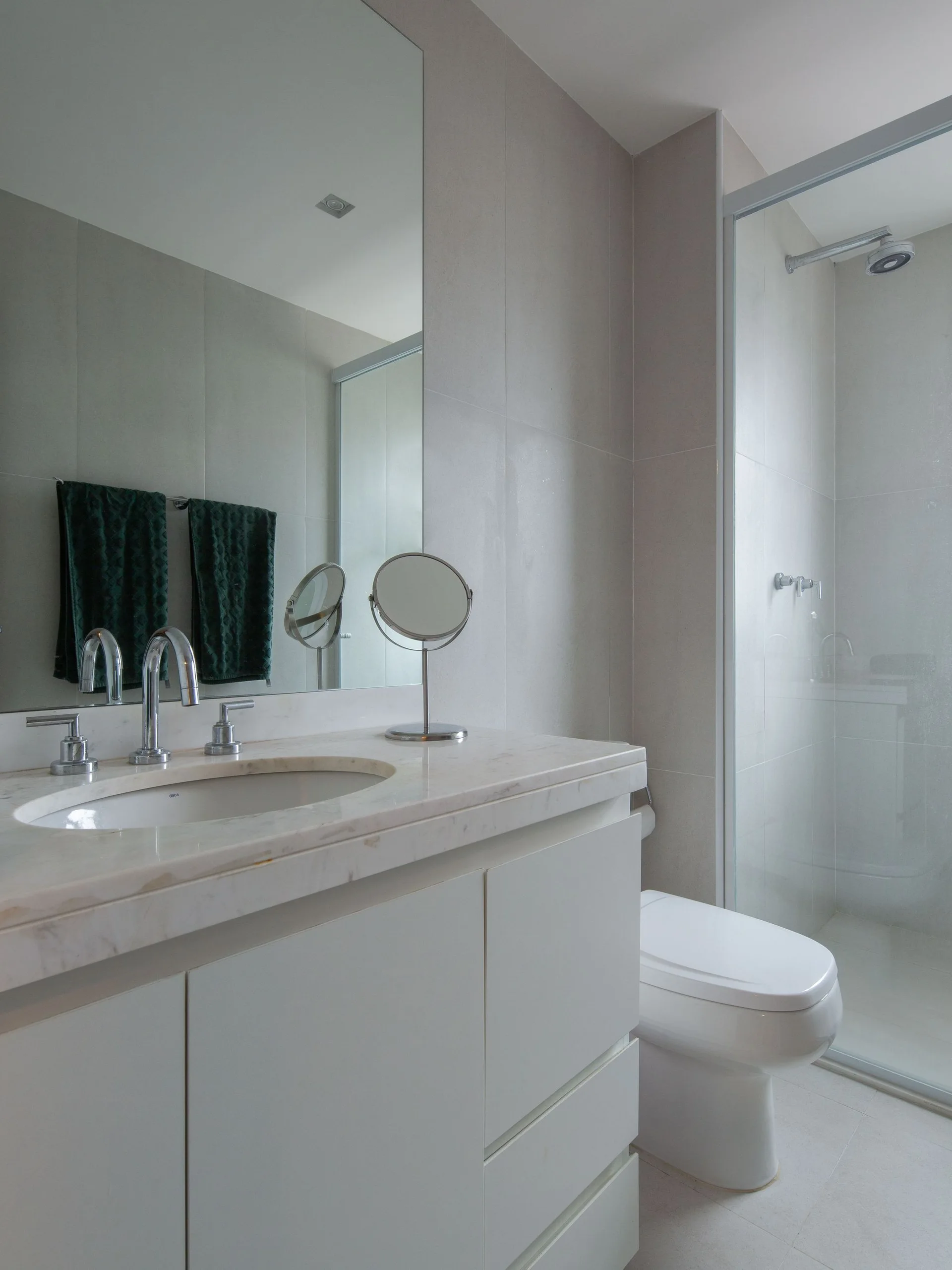 A contemporary beige bathroom features a white vanity with a stone countertop, a large rectangular mirror, a white toilet, and a glass-enclosed shower.
