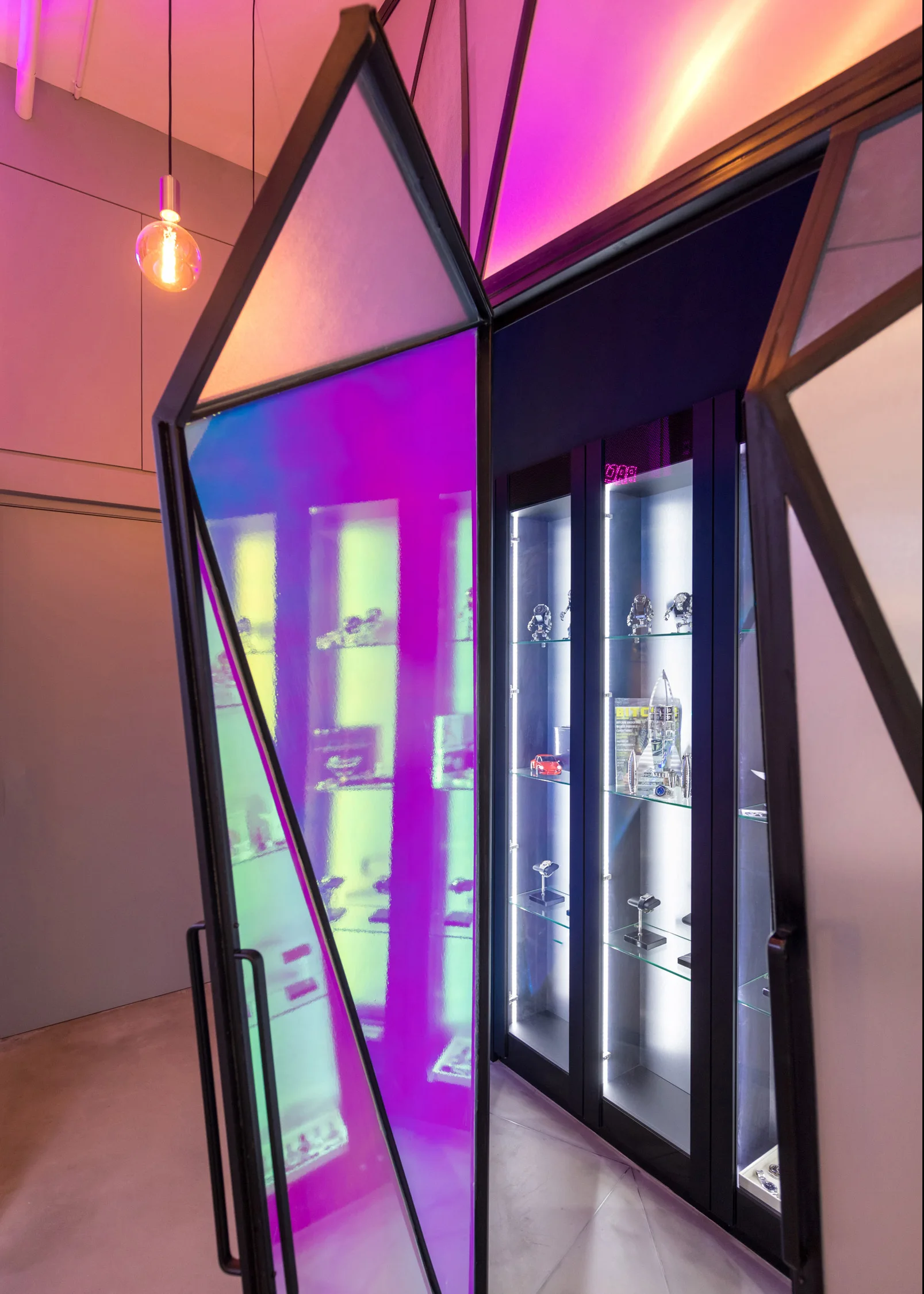 A backlit black metal display cabinet is viewed through an open geometric door featuring iridescent purple and green dichroic glass panels.