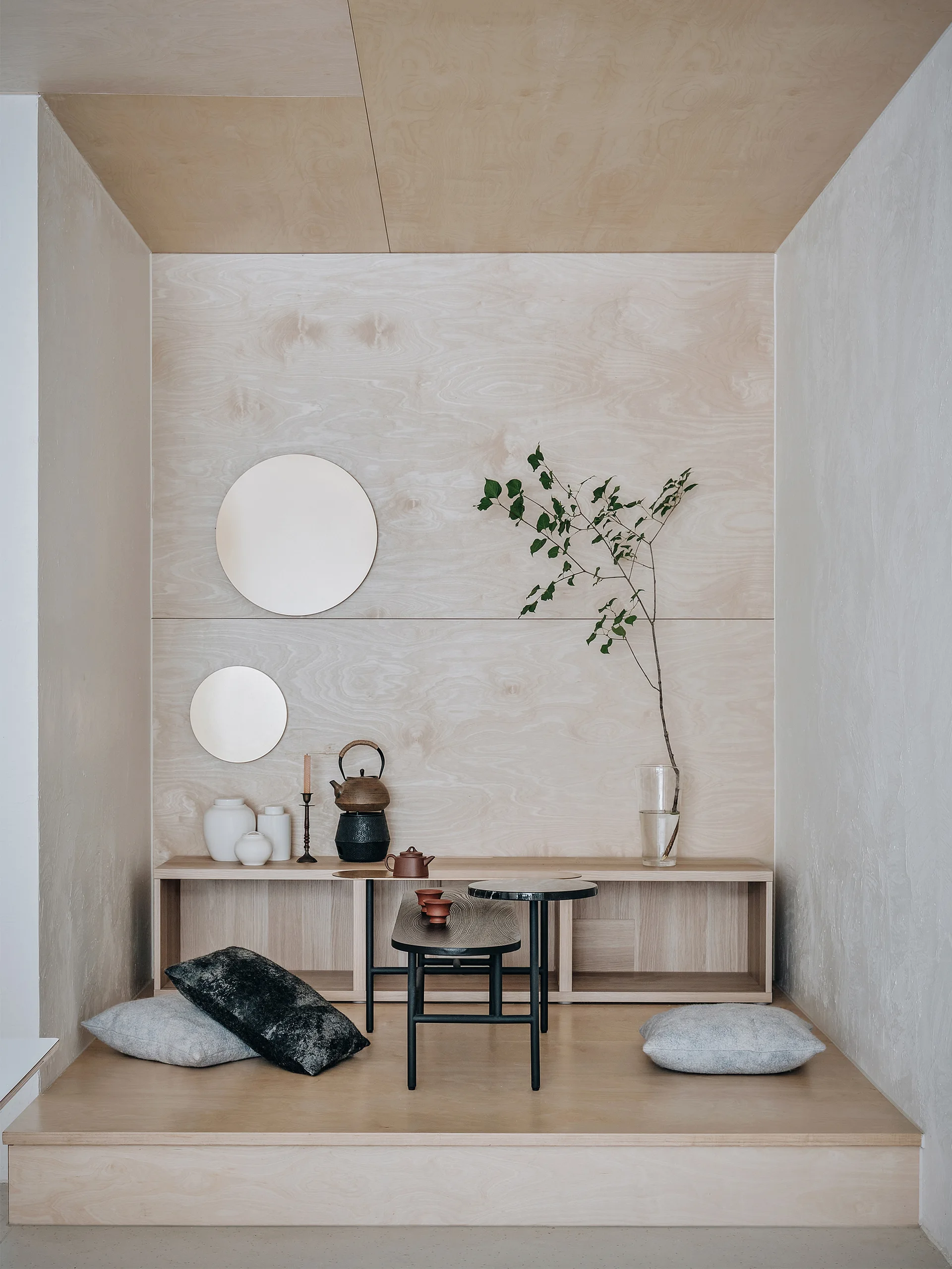A light wood paneled alcove featuring a raised platform with cushions, low wooden shelving, black tea tables, and two round wall mirrors.