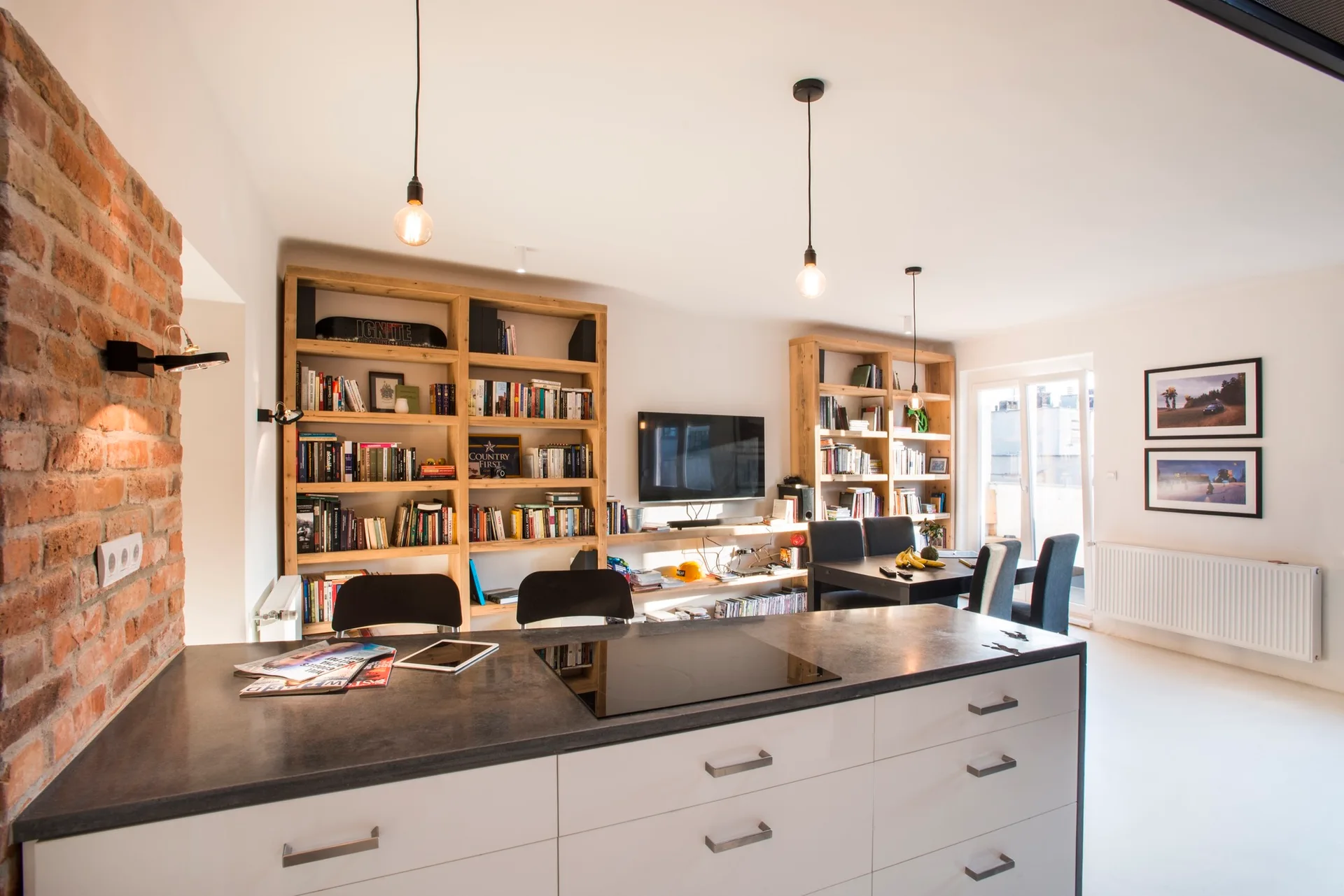A bright open-plan kitchen features a white island with a dark countertop next to an exposed brick wall, with wooden bookshelves in the background.