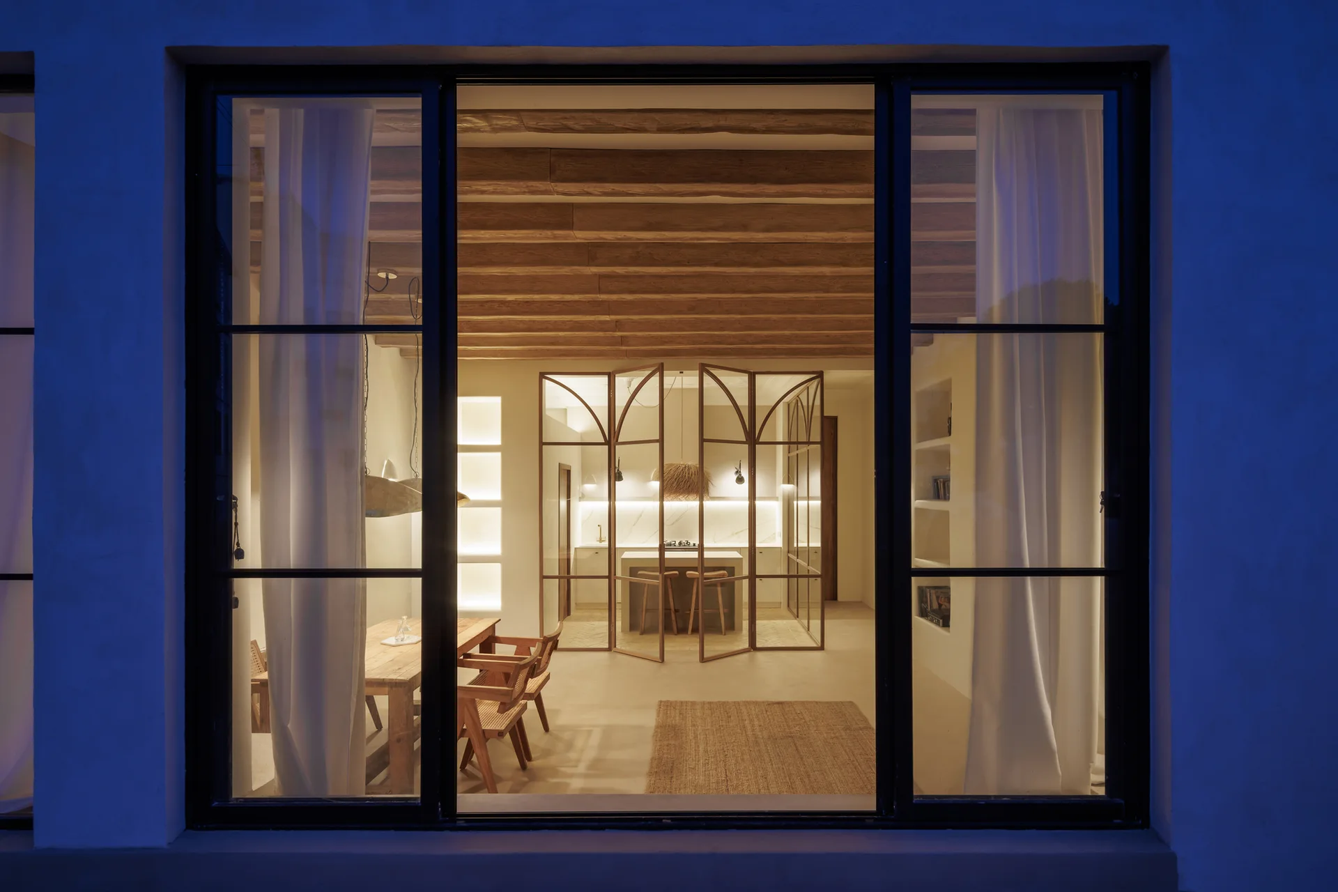 A view through large black-framed glass doors reveals a warm dining room with exposed wood ceiling beams and an arched glass kitchen divider.