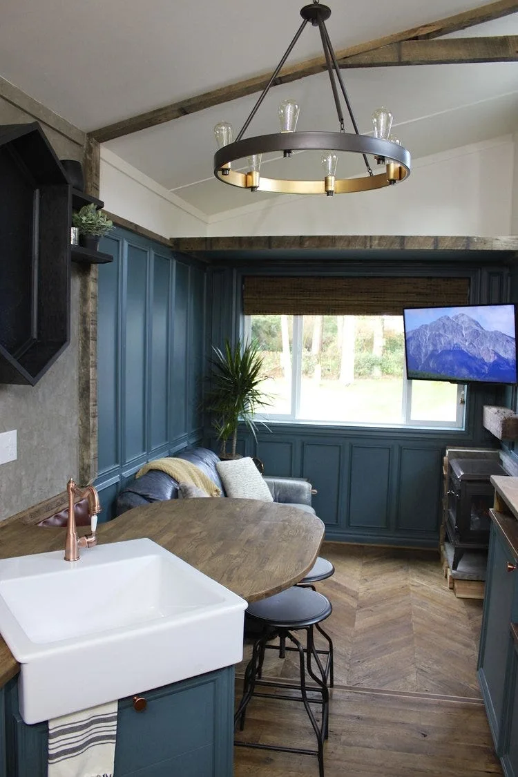 A compact blue paneled kitchen and living space features a white farmhouse sink, a curved wood peninsula counter, and chevron patterned wood flooring.