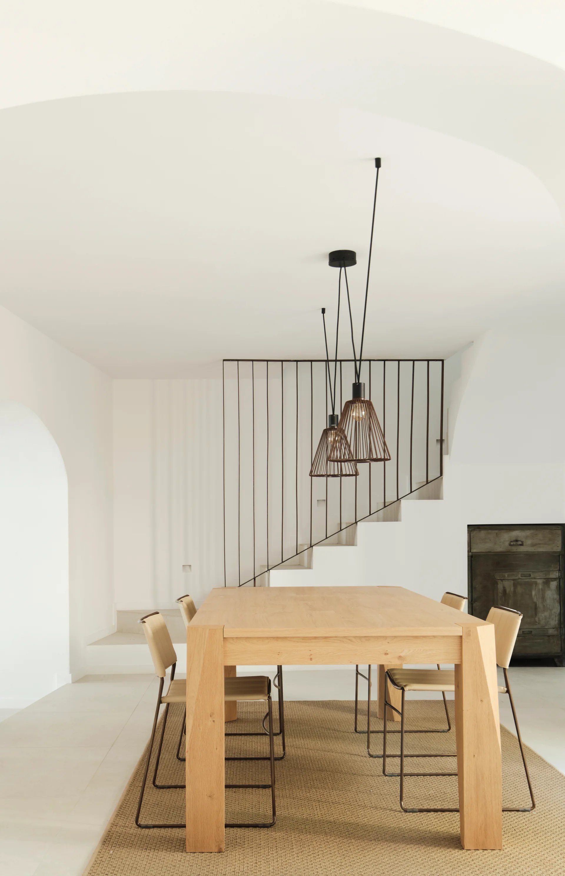 A minimalist dining room features a light wood table on a woven rug, black wire pendant lights, and a staircase with slim metal railings.