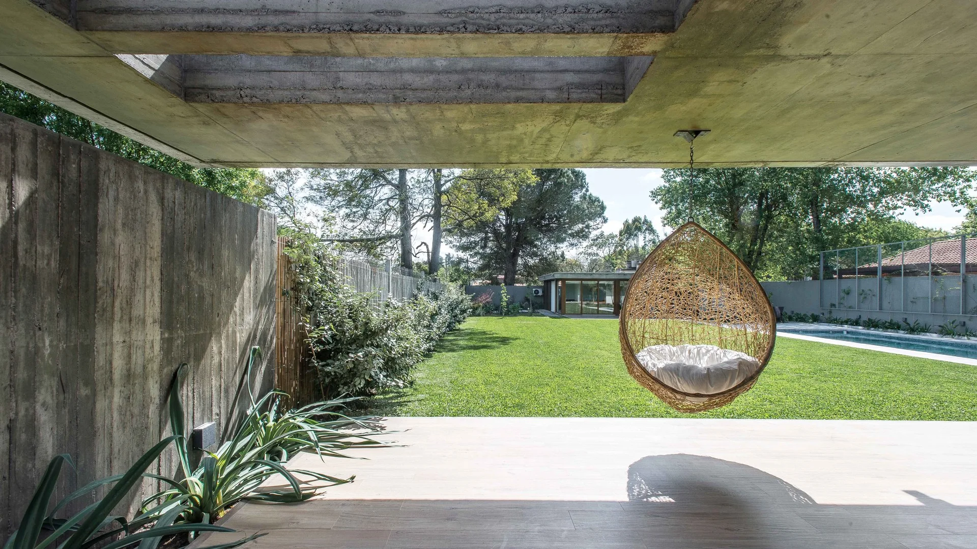 A covered concrete patio features a woven hanging egg chair with a white cushion, overlooking a large green lawn and swimming pool.