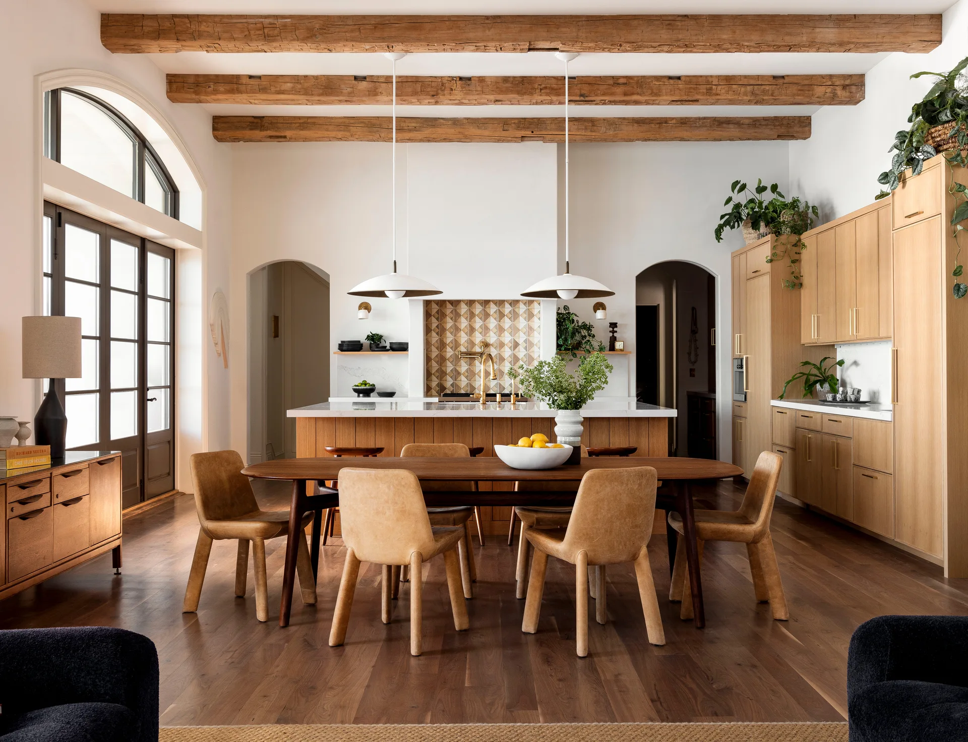A bright open-concept kitchen and dining area with exposed wood ceiling beams, tan leather dining chairs, and warm wood cabinetry.