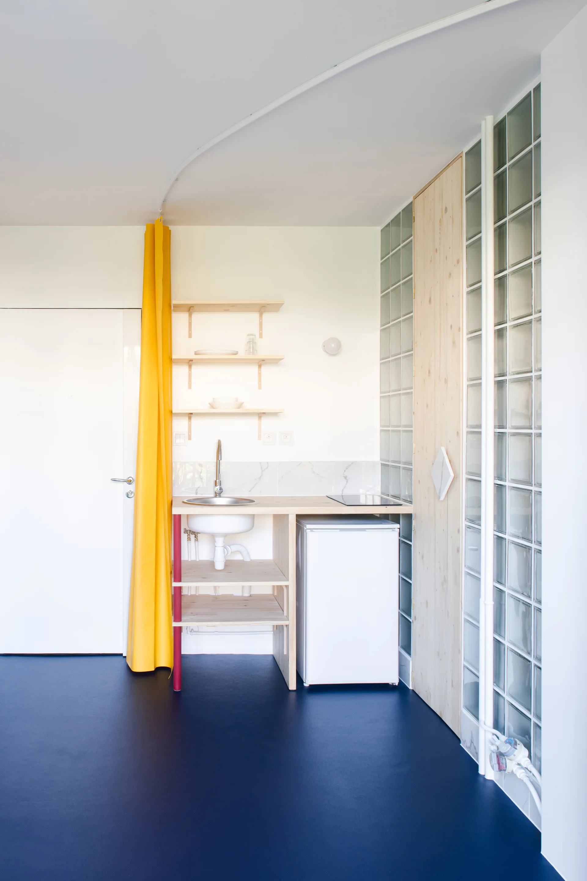 A compact kitchenette with a light wood counter, under-counter fridge, open wooden shelves, a bright yellow curtain, and a glass block wall.