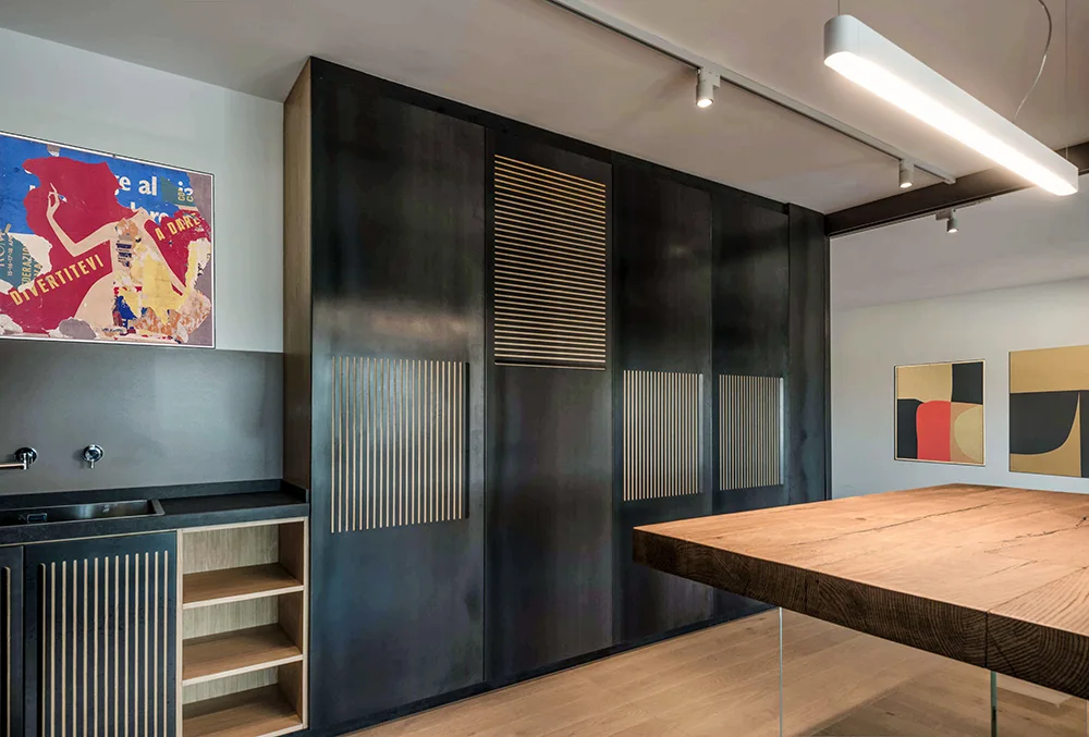 A modern kitchen features full-height dark gray cabinets with slatted wood details, a thick wooden island table, and a linear pendant light.