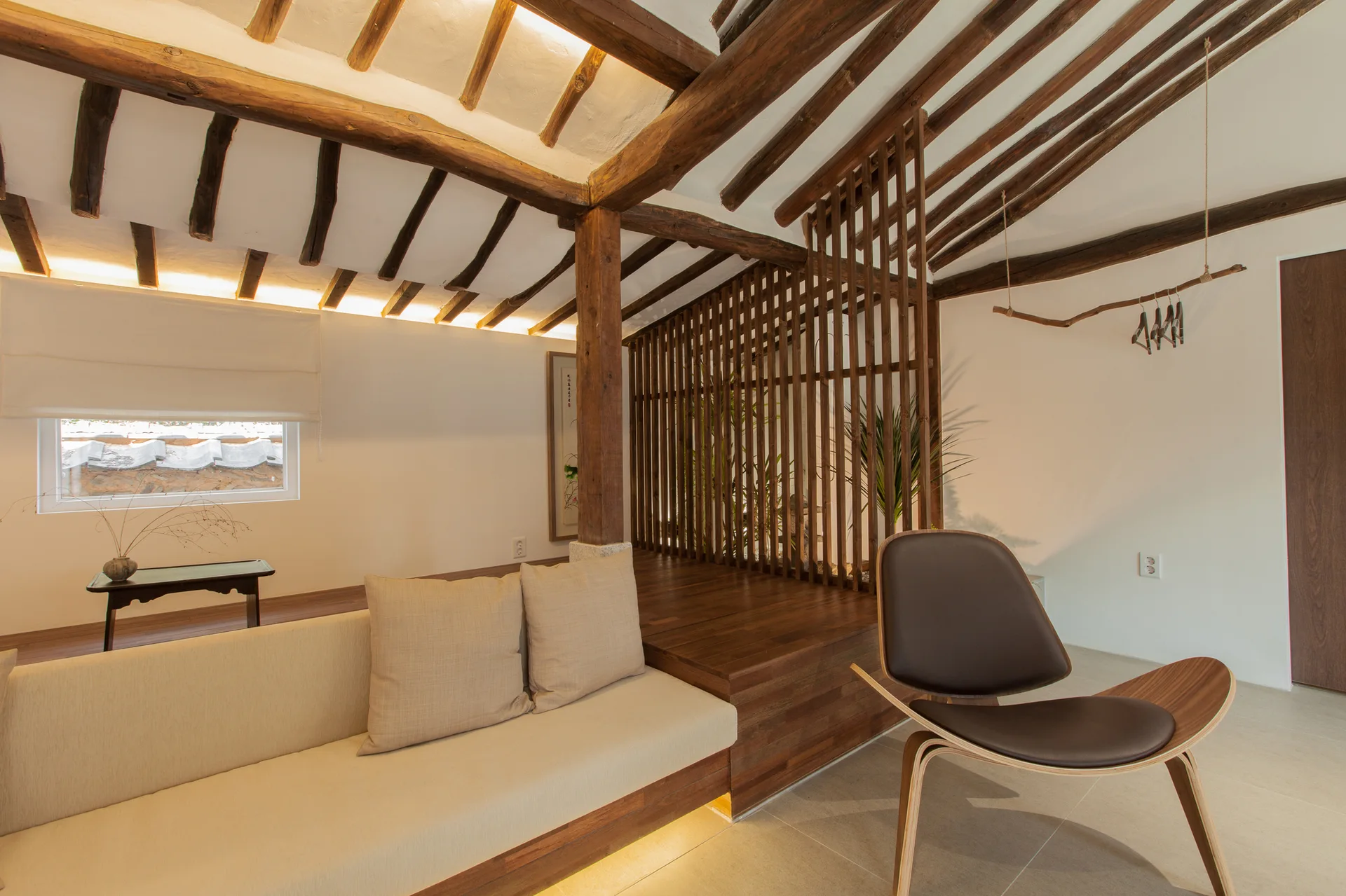 A minimalist living area shows exposed dark timber ceiling beams, a built-in platform sofa with lighting below, and a wooden slat partition.
