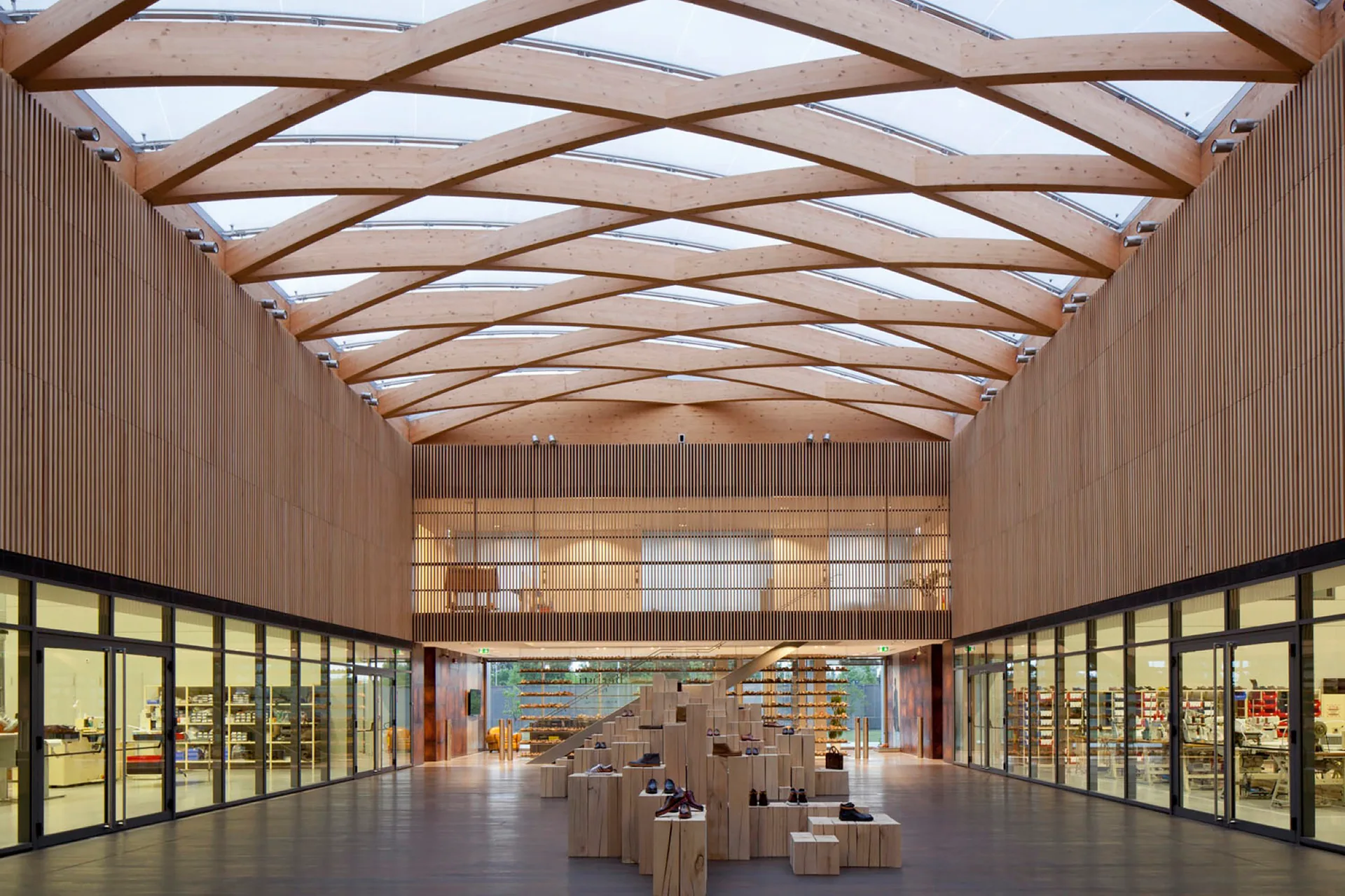 A spacious atrium with a crisscrossing wood beam ceiling, vertical timber slat walls, glass partitions, and a central tiered wooden block display.