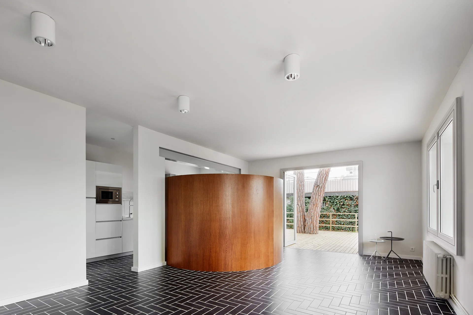 A minimalist room with a large curved wooden partition, dark herringbone floor tiles, white walls, and a door opening to a patio.
