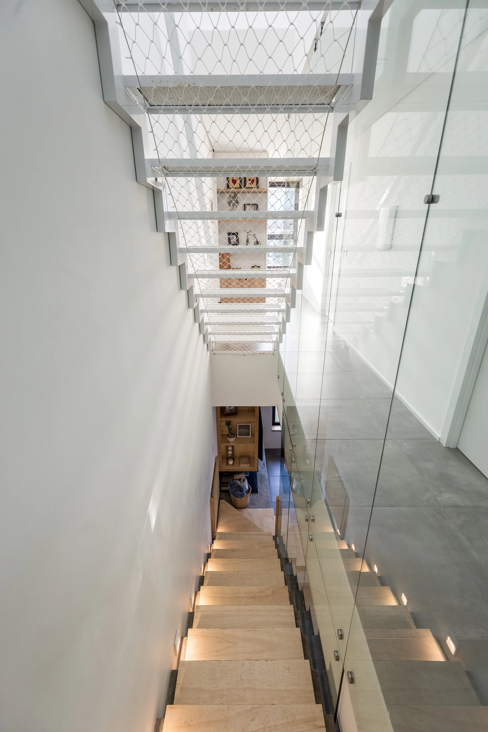 A modern staircase features light wood steps with integrated lighting, a transparent glass railing, and a white wire mesh partition overhead.