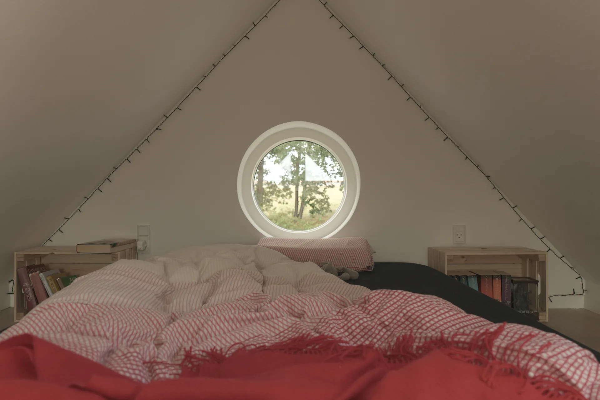 A low bed with red and white bedding sits under a sloped attic ceiling with a circular window, flanked by wooden crate nightstands.