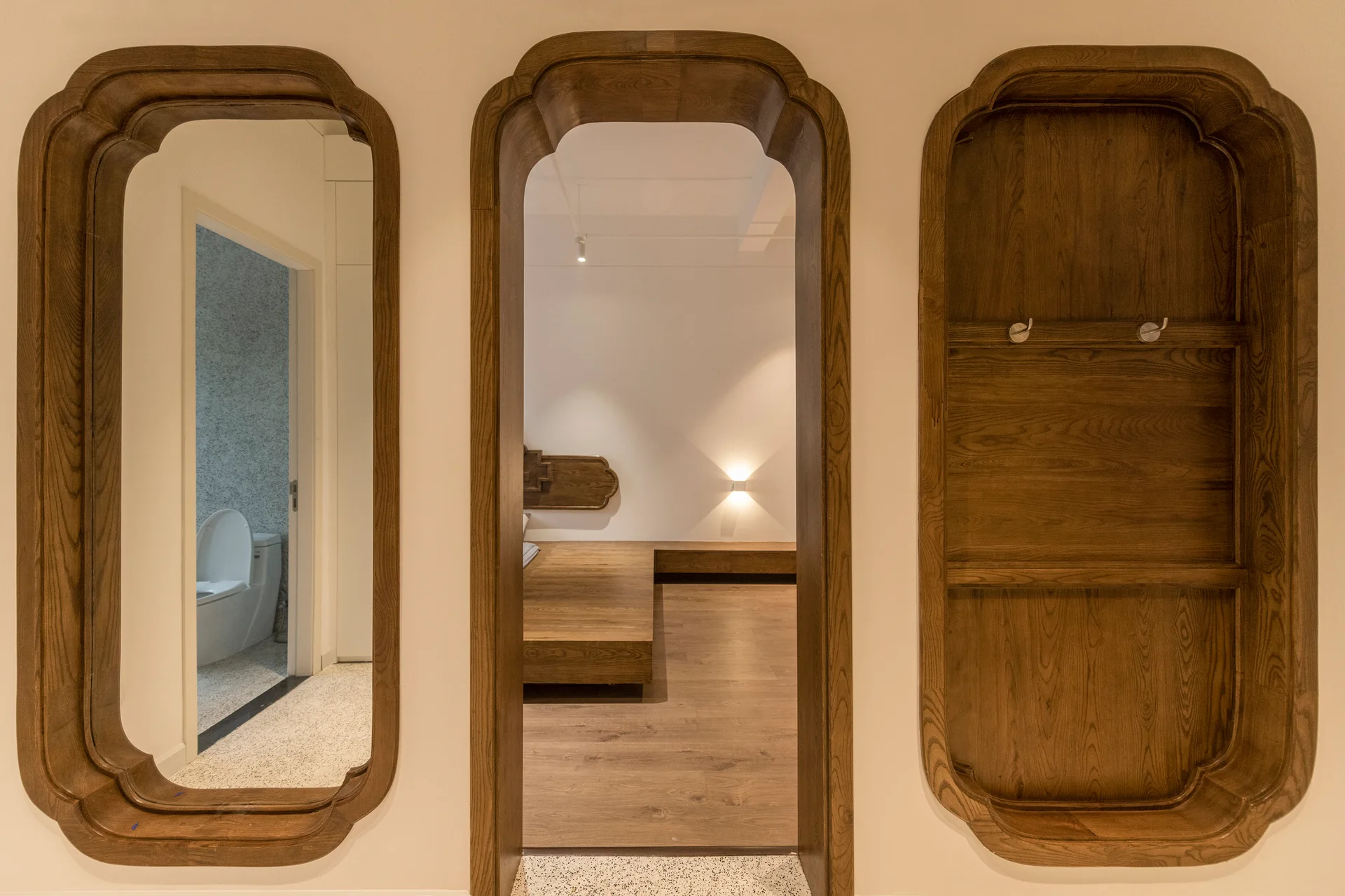 Three matching rounded wooden frames set into a light cream wall house a mirror, an open bedroom doorway, and a recessed shelving niche.