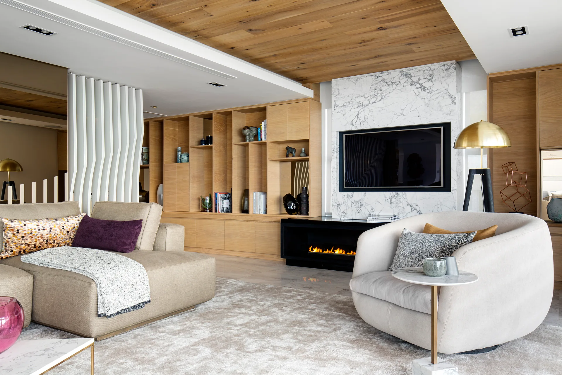A contemporary living room features a beige sectional, a curved armchair, light wood built-ins, and a veined marble fireplace surround.