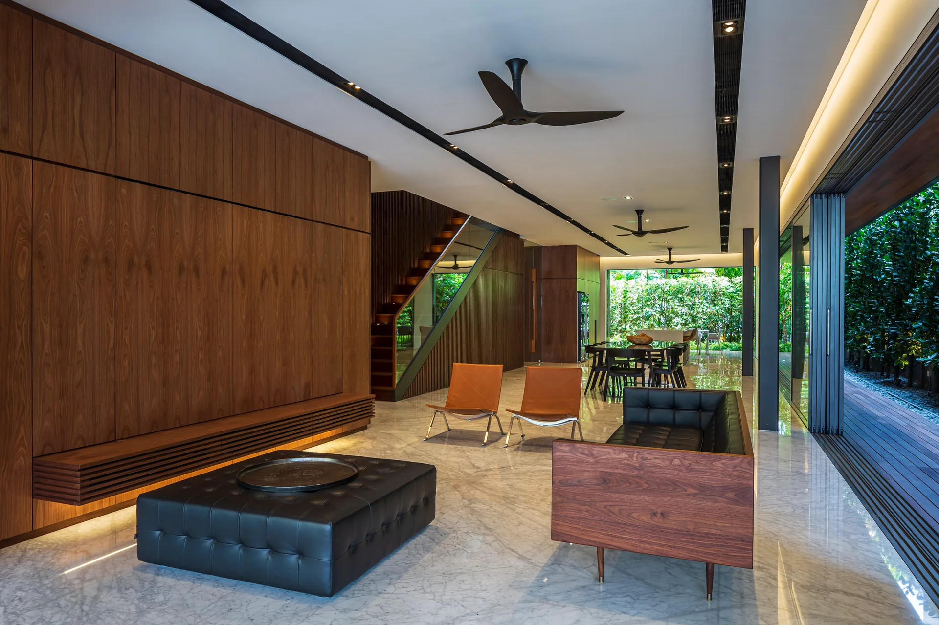 A modern open-plan living room with walnut paneled walls, black leather seating, polished marble floors, and large sliding glass doors.