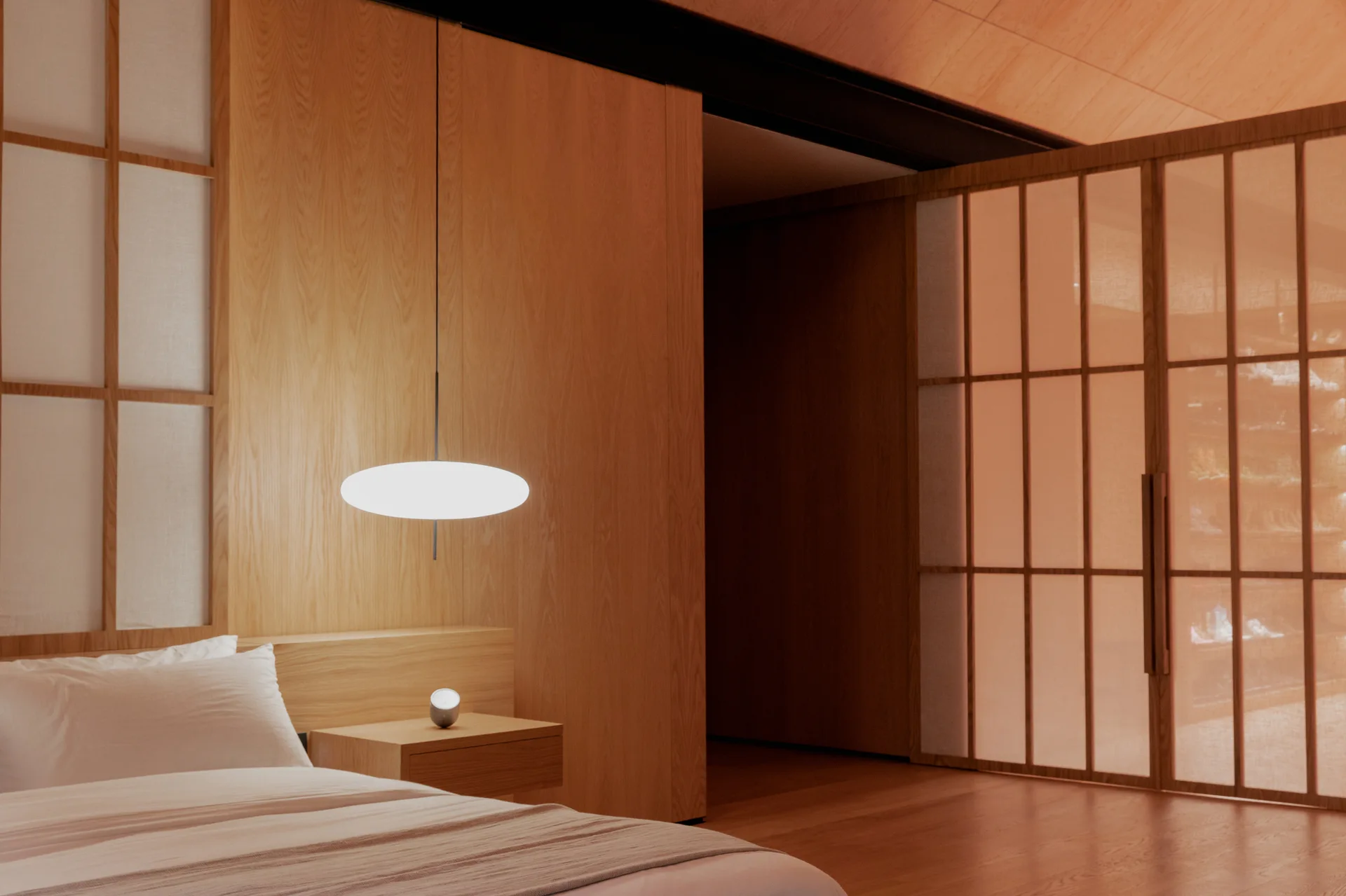 A minimalist bedroom featuring warm wood paneled walls, a low bed with white bedding, a floating wooden nightstand, and sliding shoji screens.