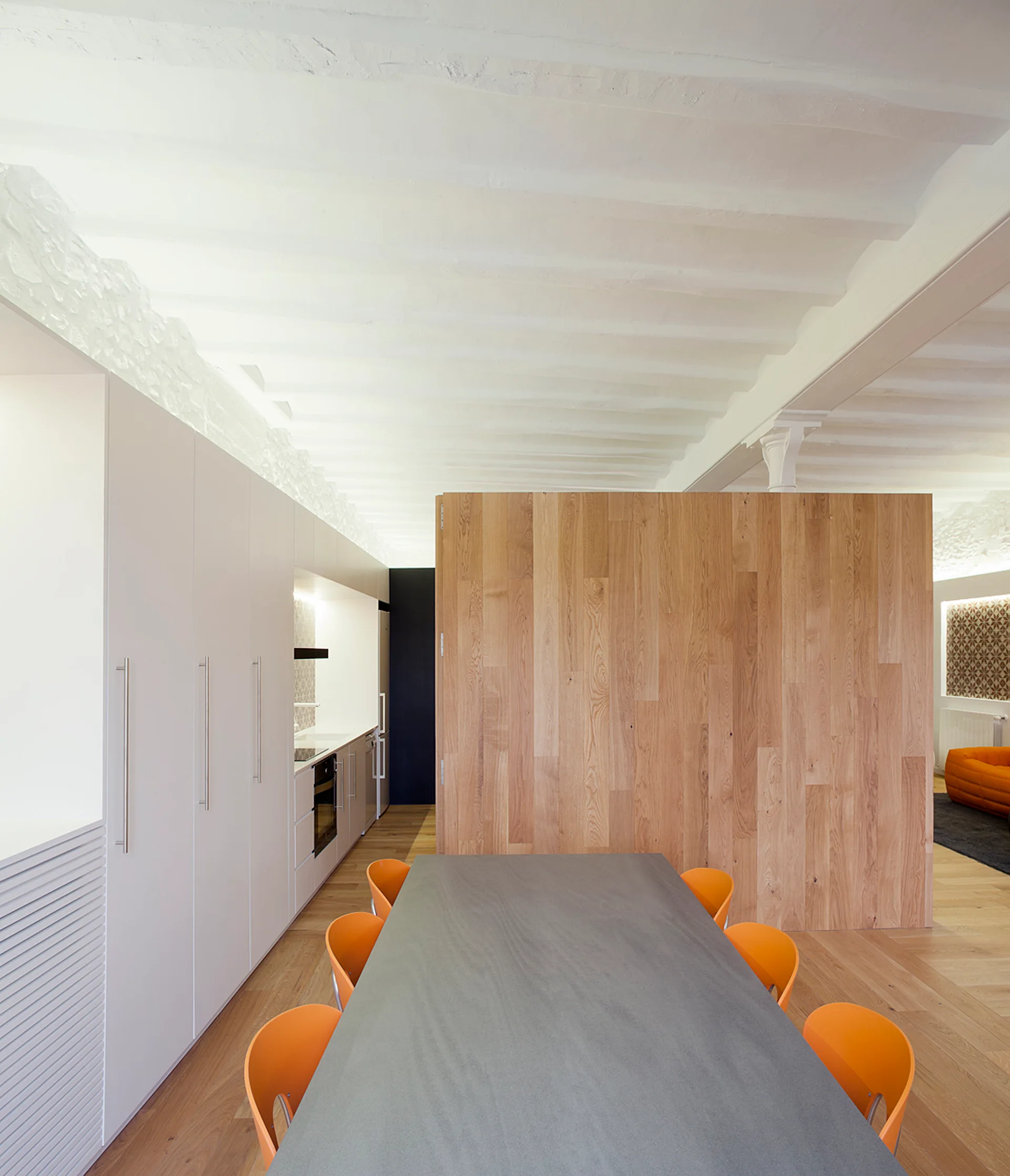 A grey dining table with orange chairs sits near a large wood-paneled partition wall and flat-front white kitchen cabinets under a beamed ceiling.