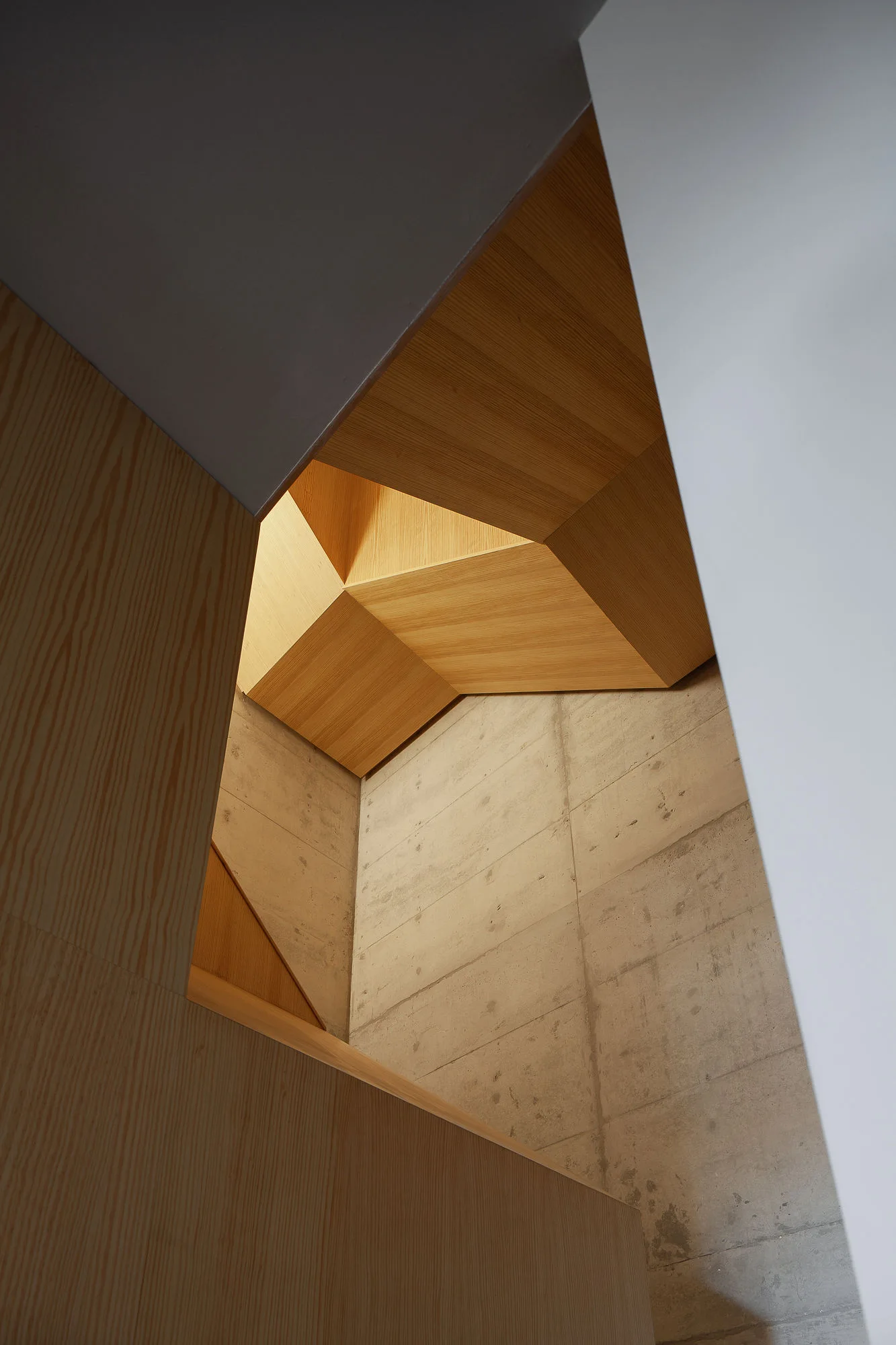 An upward view of a geometric light wood soffit ceiling integrated with an exposed grey concrete wall inside a minimalist stairwell.