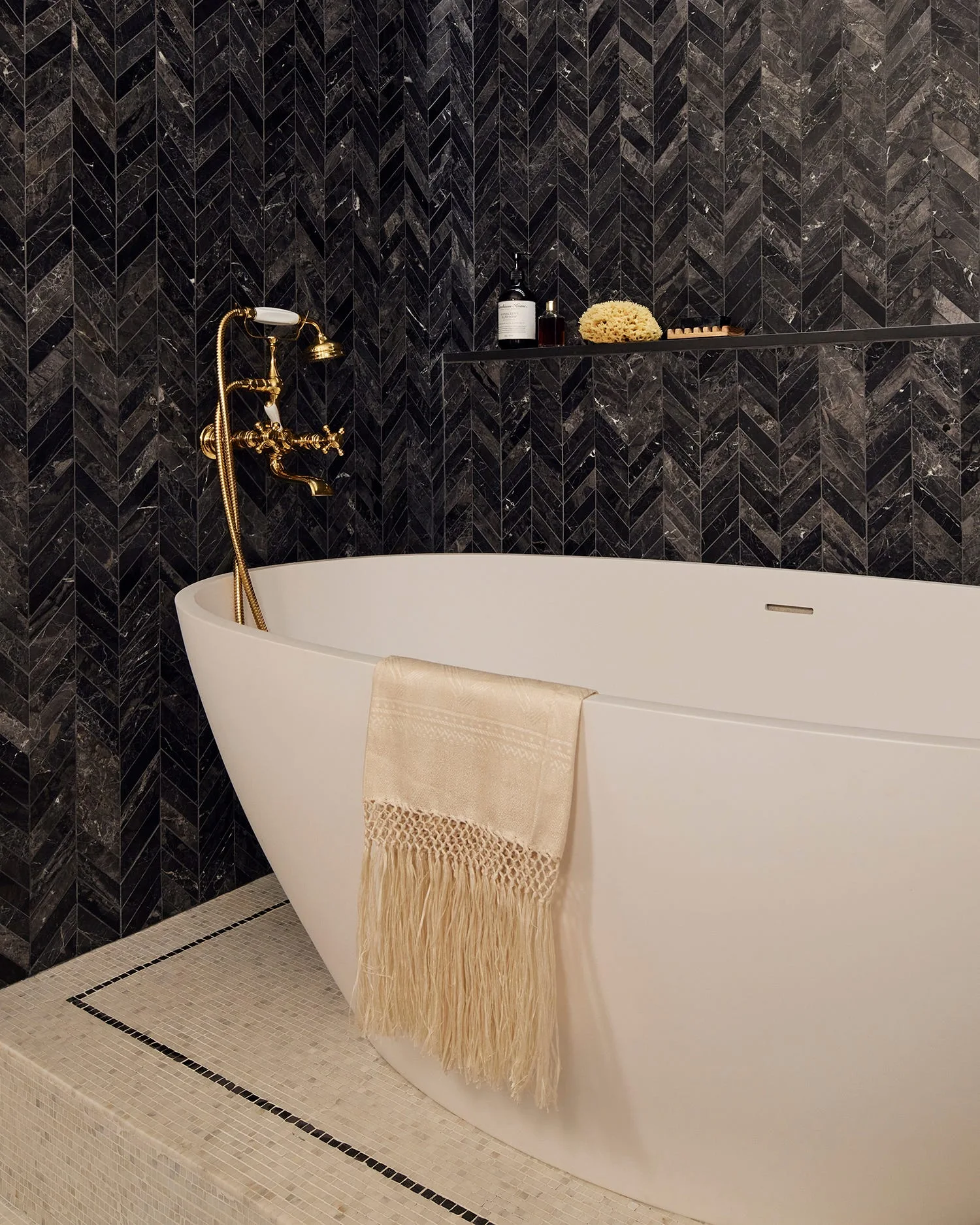 A white freestanding bathtub features a gold faucet and a draped cream towel against a black herringbone tile accent wall.