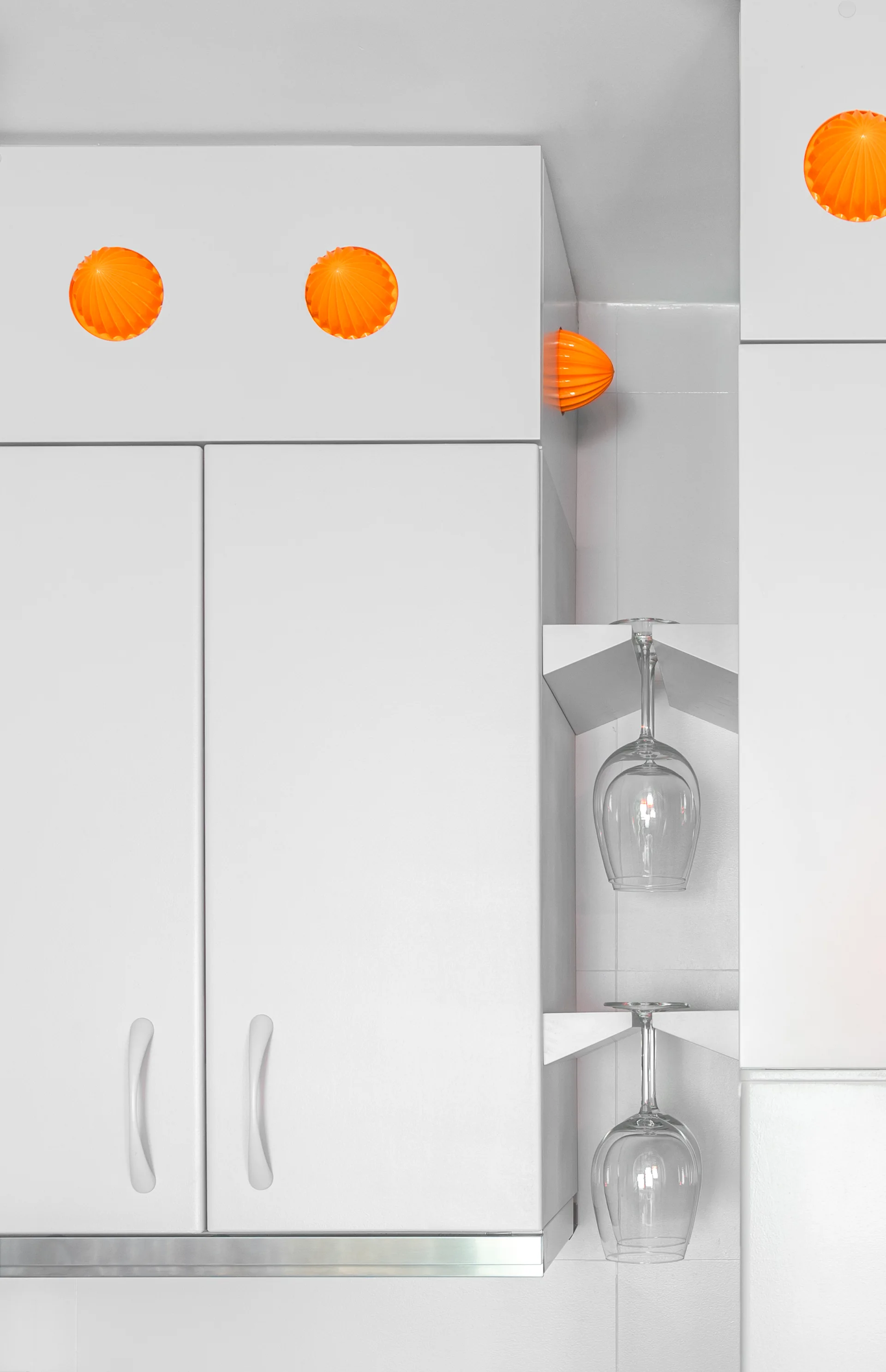 Matte white kitchen cabinets feature bright orange circular knobs and a vertical open shelf housing upside-down wine glasses.