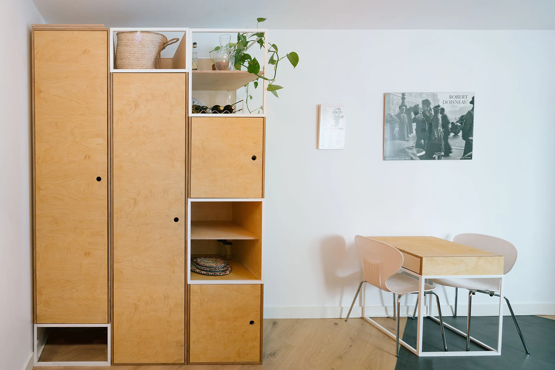 A minimalist room features a tall custom plywood storage unit next to a small wooden dining table with white chairs against a white wall.
