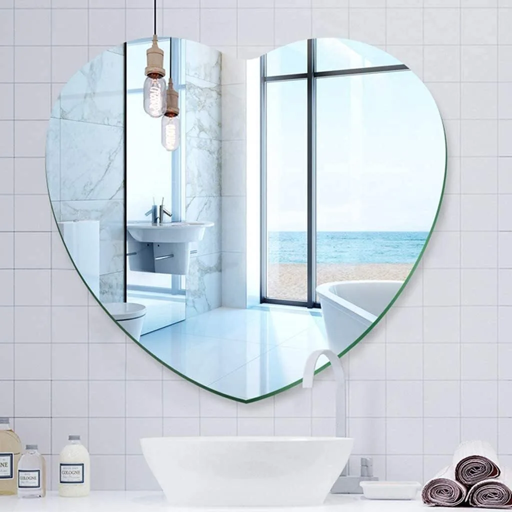 A frameless heart-shaped mirror hangs on a white grid-tiled wall above a white vessel sink, reflecting a bright bathroom with an ocean view.