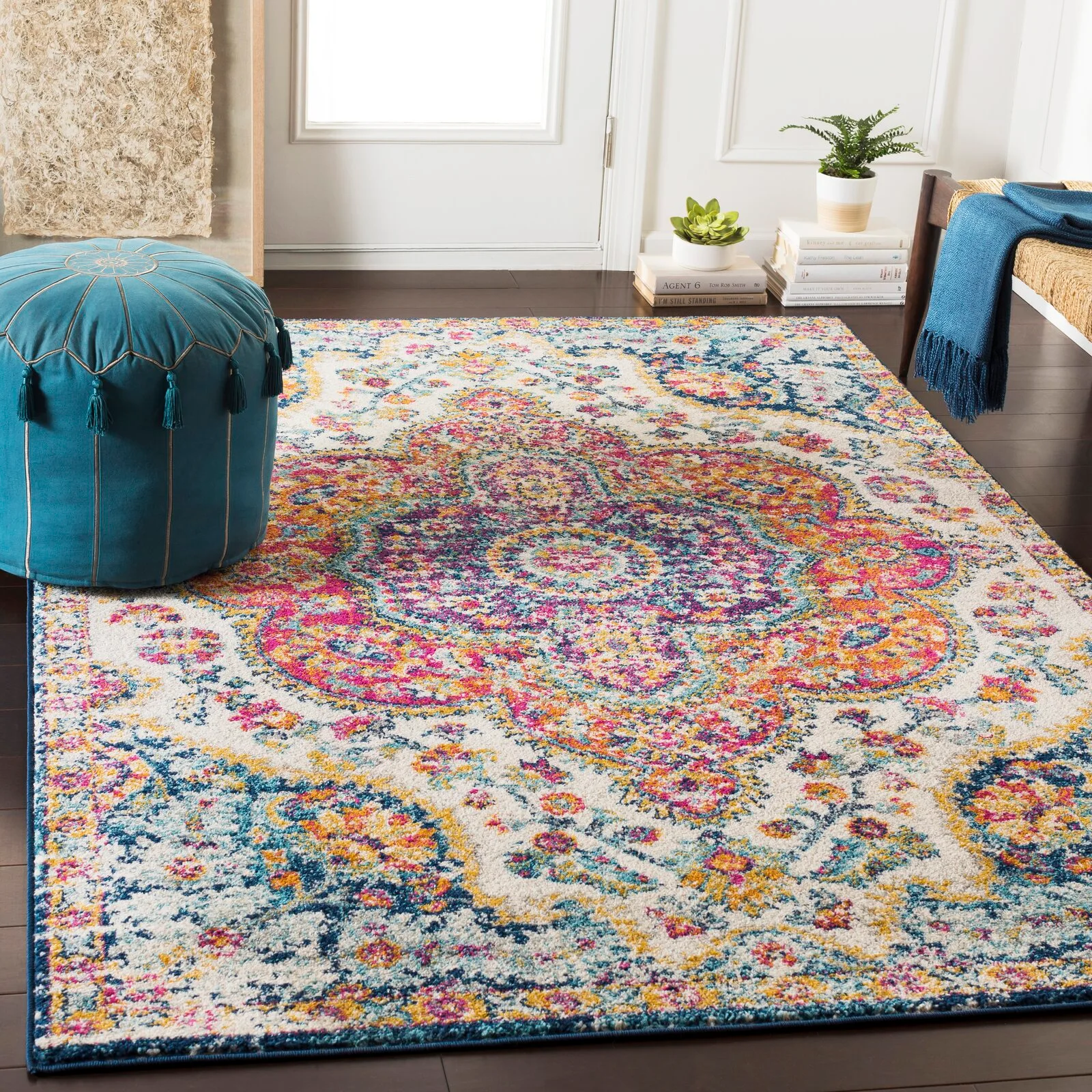 A colorful medallion area rug with pink, blue, and yellow patterns rests on a dark hardwood floor next to a teal tasseled ottoman.
