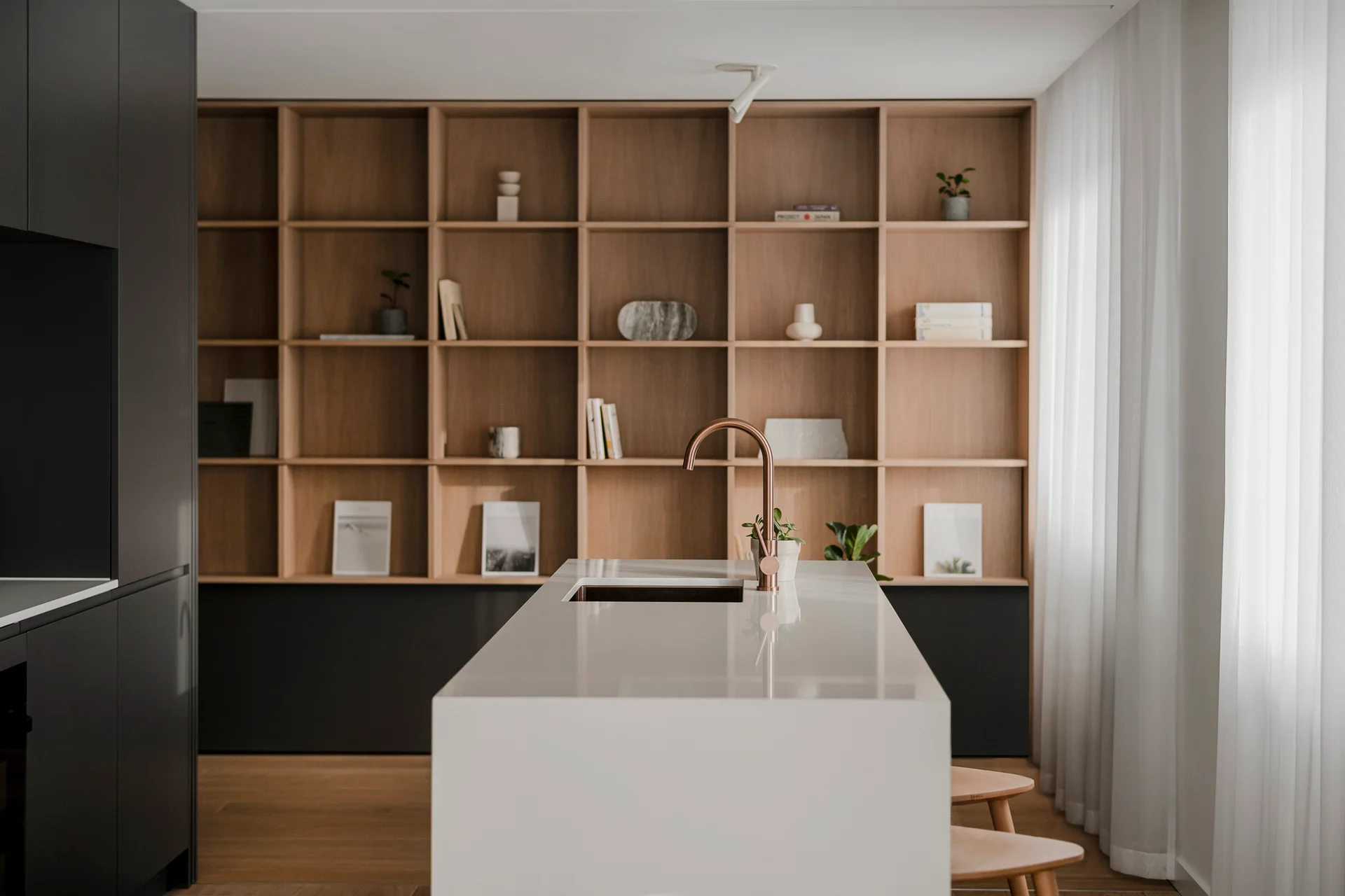 A modern kitchen featuring a sleek white island with a copper faucet, positioned in front of a floor-to-ceiling light wood grid shelving unit.