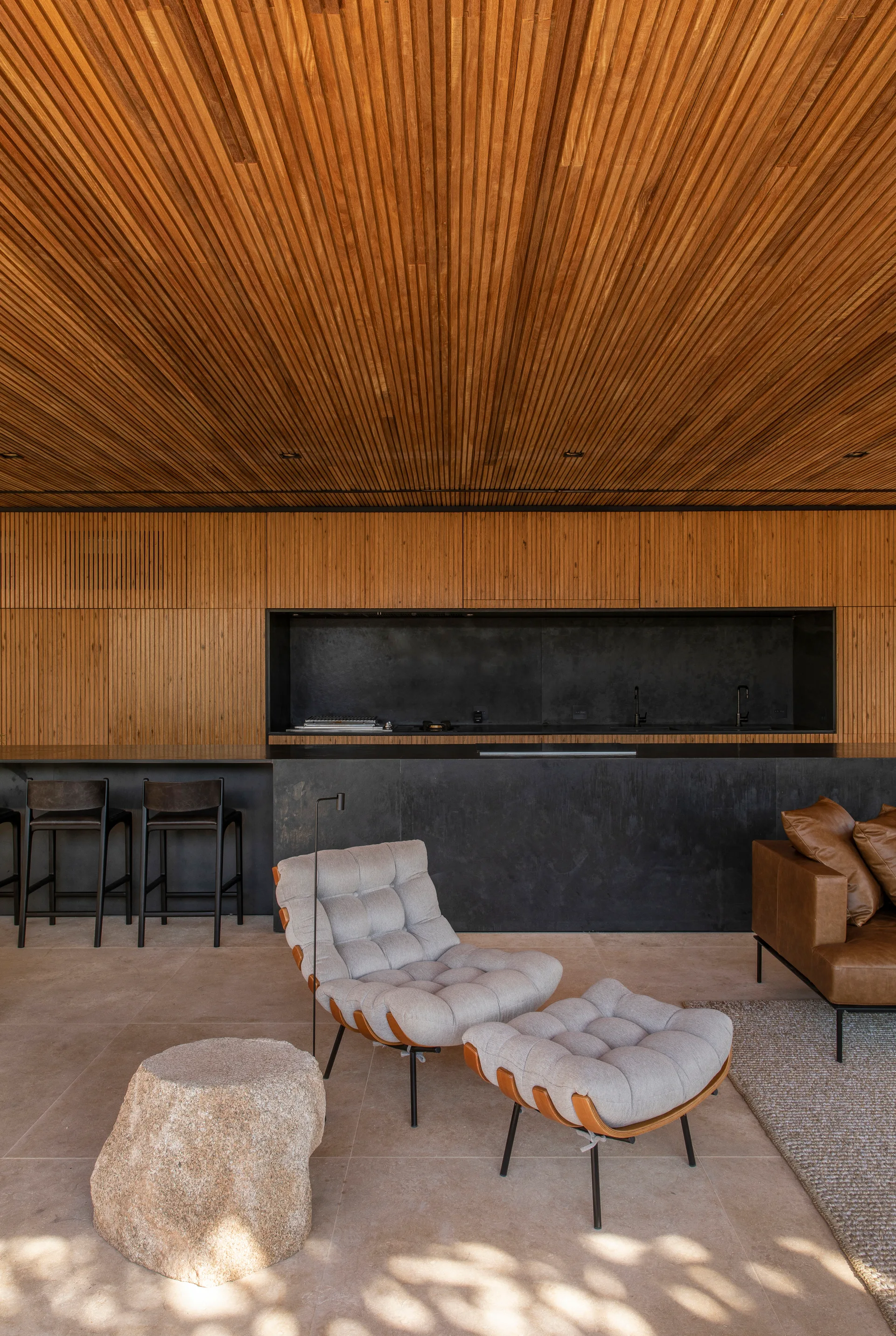 A modern living area features a slatted wood ceiling, a tufted grey lounge chair with ottoman, a raw stone side table, and a black kitchen in the background.
