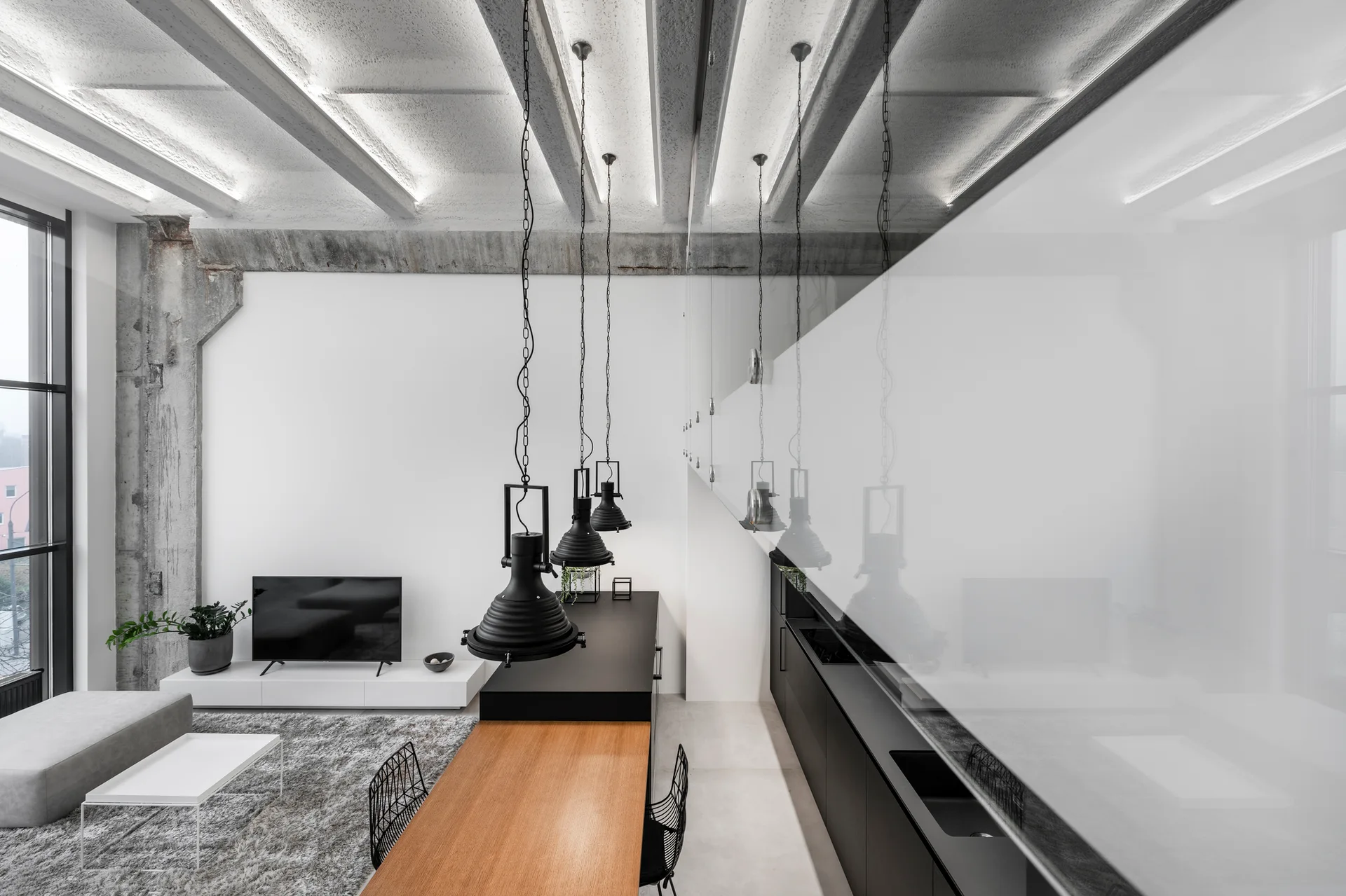 A modern open-plan loft featuring exposed concrete ceiling beams, a long wooden dining table with black pendant lights, and a minimalist living area.