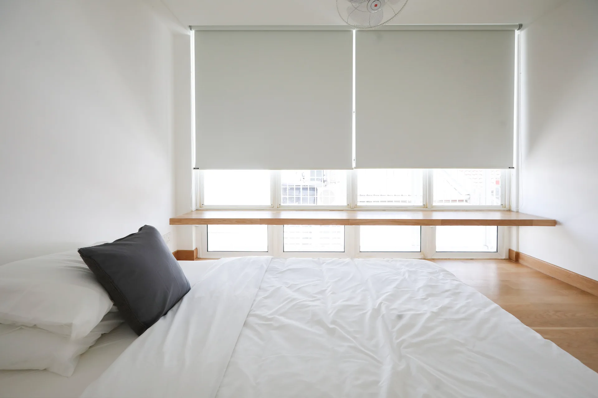 A minimalist bedroom features a low bed with white bedding next to a large window with white roller blinds and a long wooden ledge.