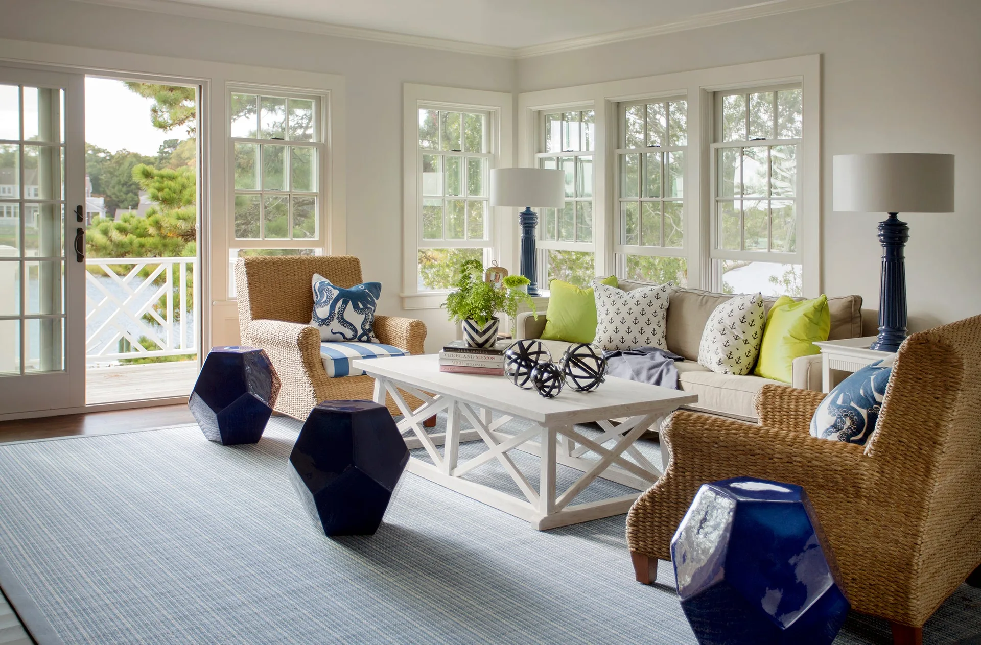 A coastal living room features a beige sofa, two wicker armchairs, three blue geometric ceramic stools, and a white coffee table on a striped rug.