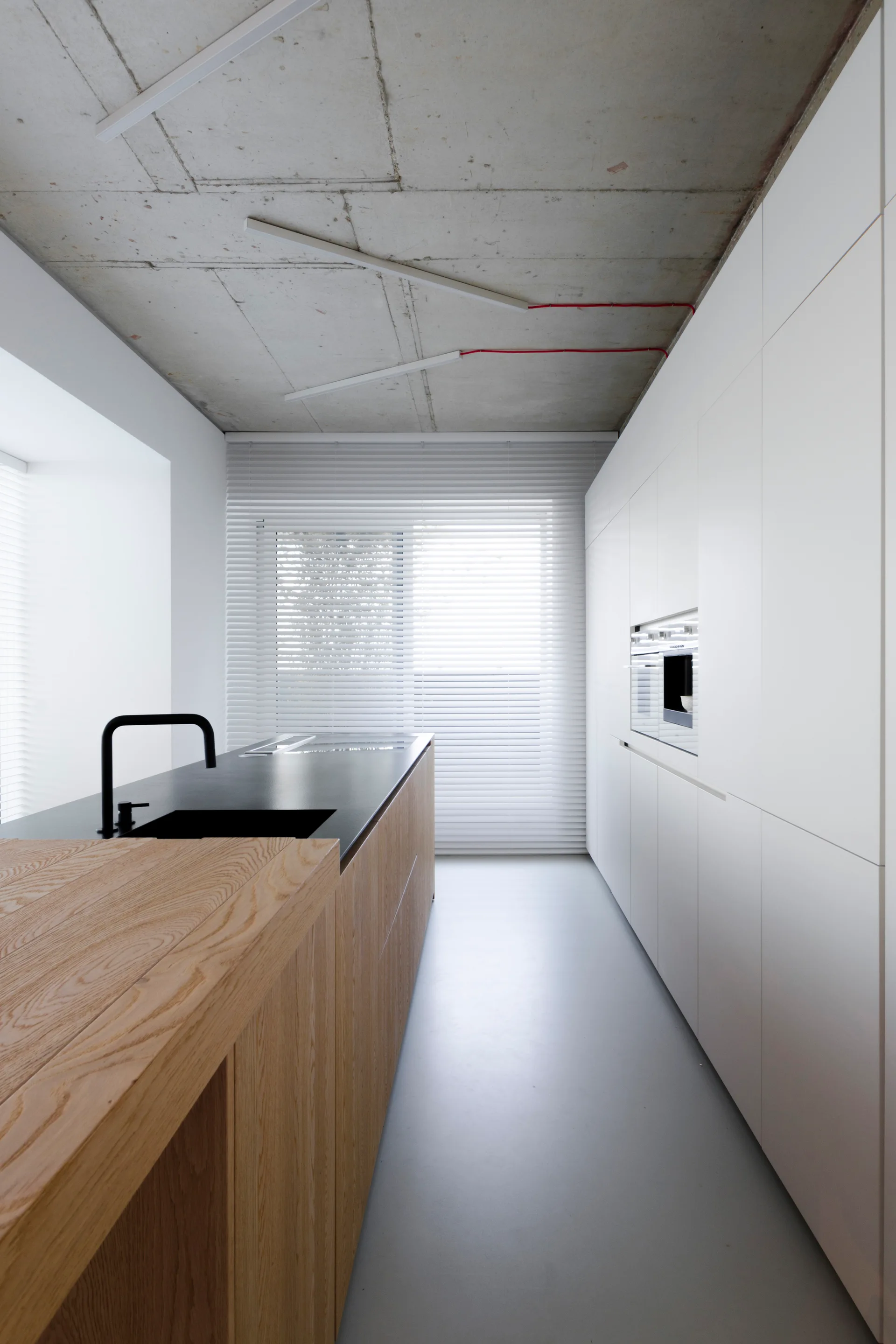A minimalist kitchen features a wood-fronted island with a black faucet, white handleless cabinetry, and an exposed raw concrete ceiling.