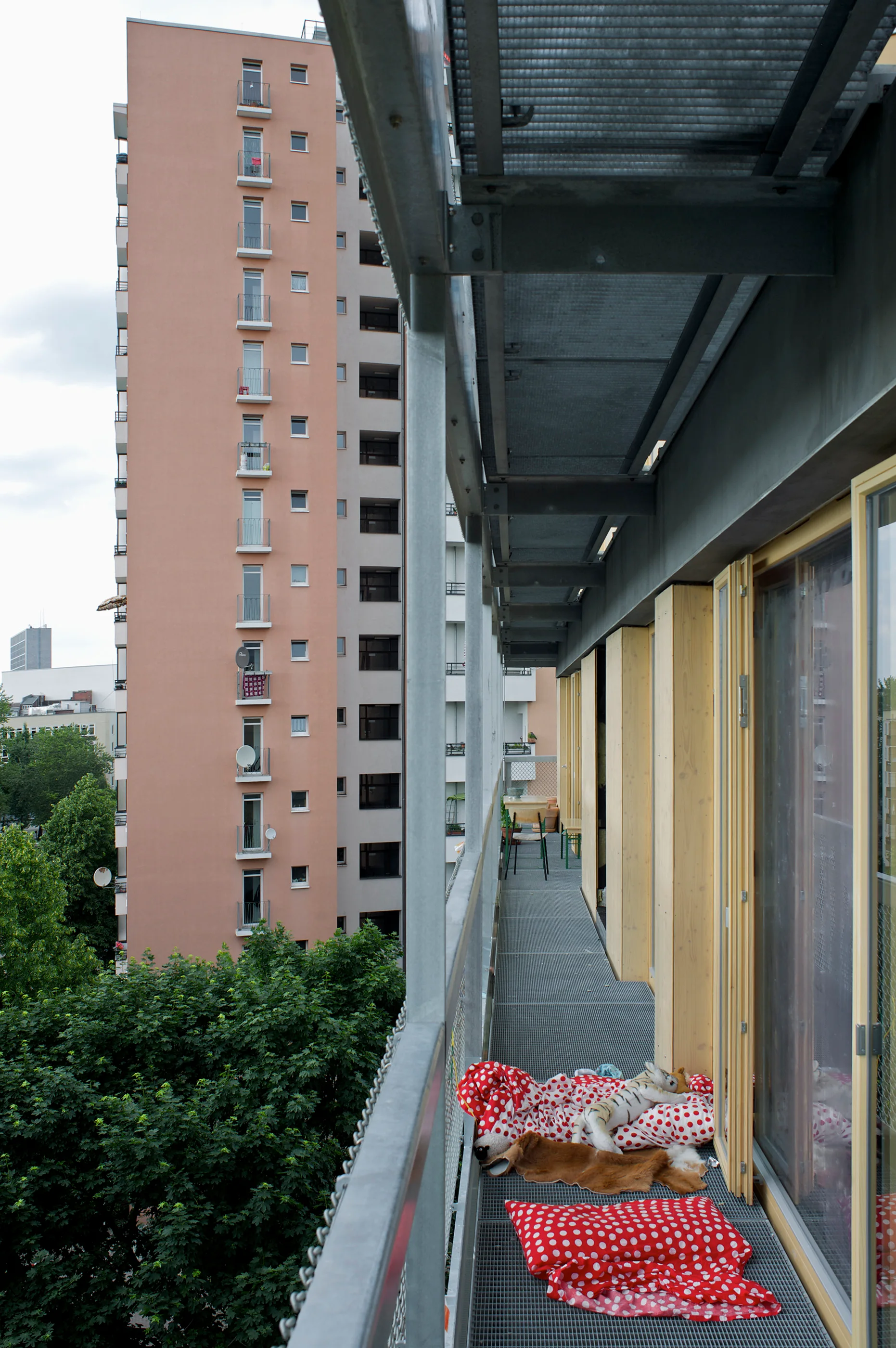 A long metal-grate balcony walkway features red and white polka dot pillows on the floor, set against a tall peach-colored residential building.