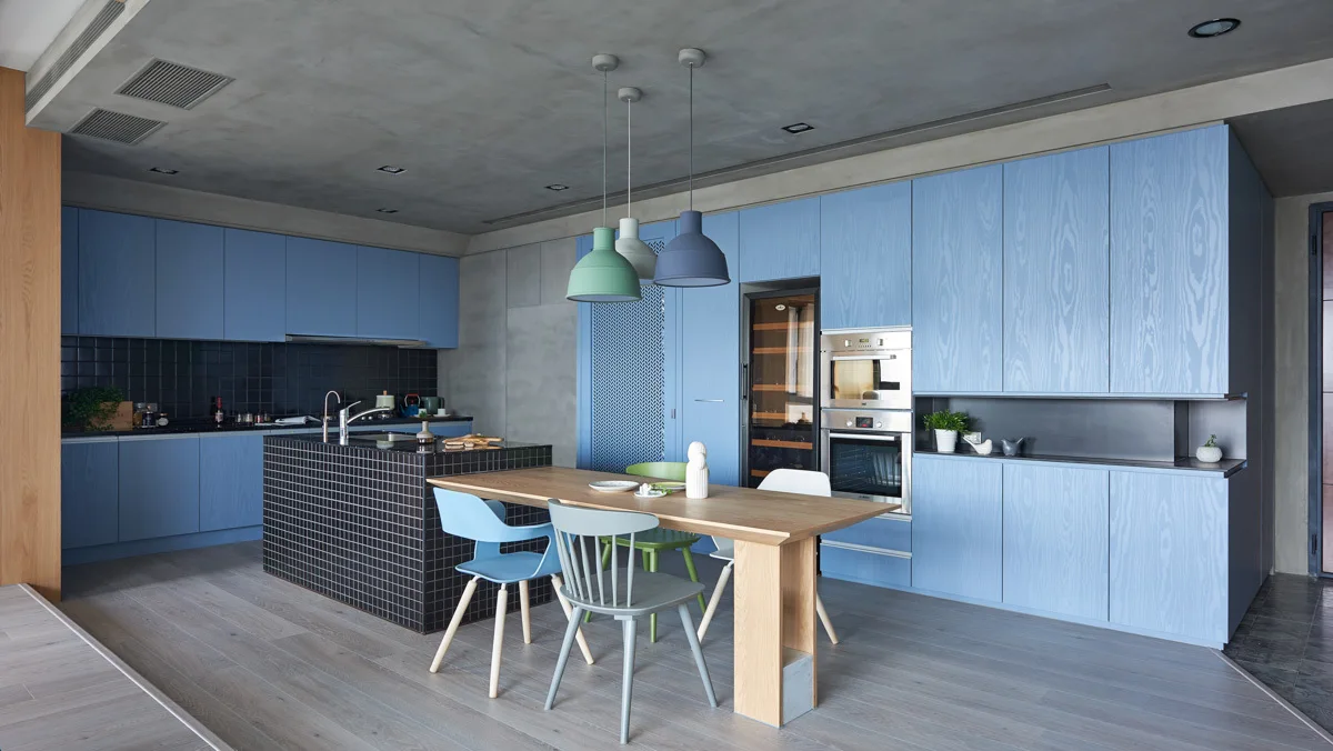 A modern kitchen with blue wood-grain cabinets, a black tiled island with an attached wooden dining table, and a grey concrete ceiling.