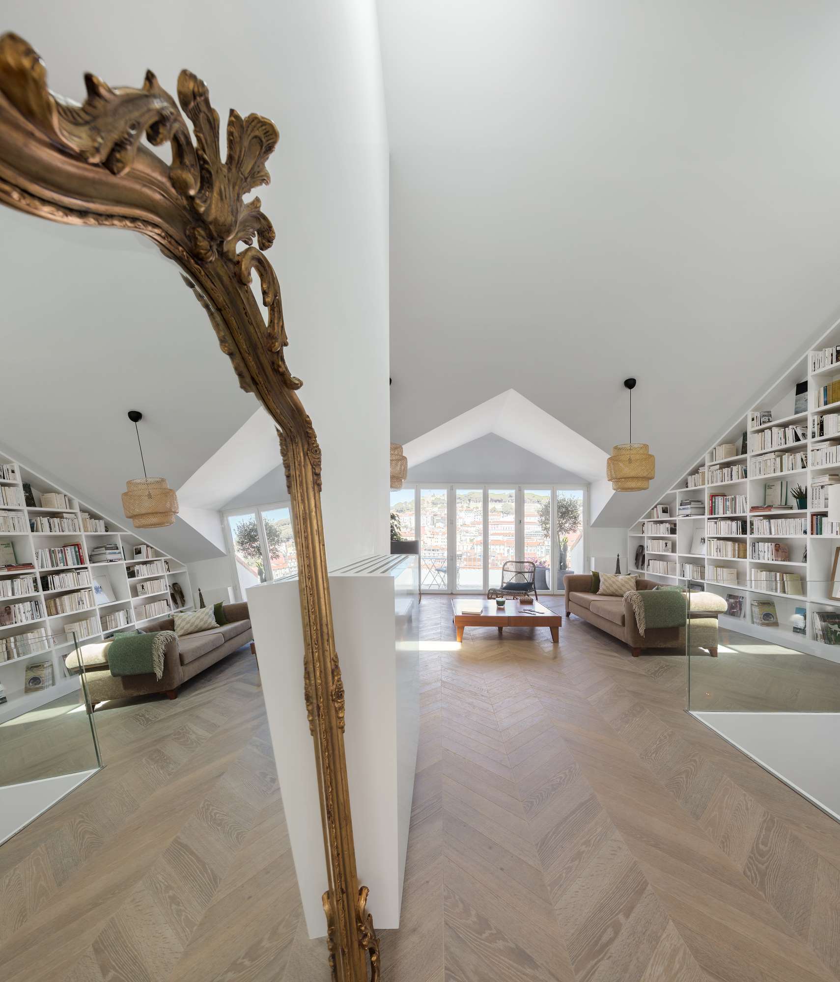A bright attic living room featuring a pitched ceiling, custom white bookshelves matching the roofline, herringbone wood floors, and a large ornate gold mirror.