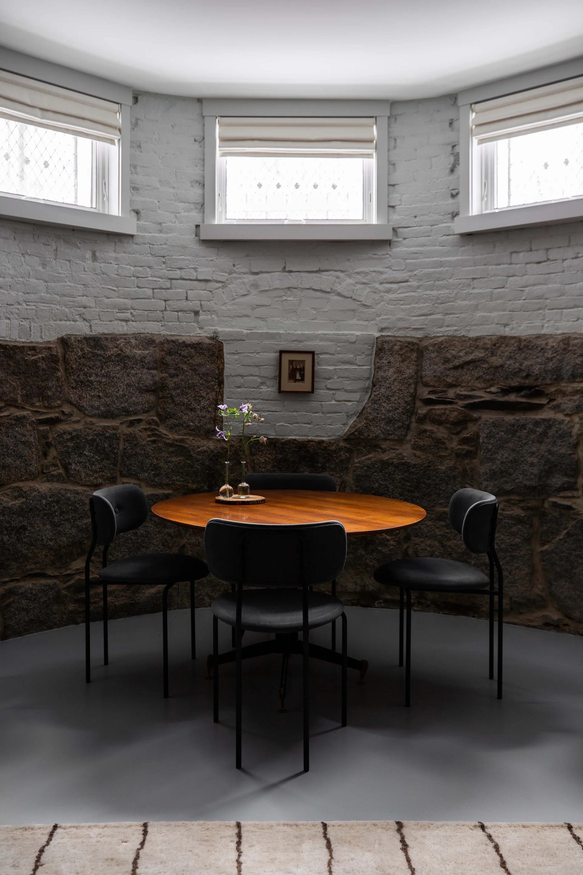 A round wooden dining table with four black chairs sits in a curved nook featuring a rough stone lower wall and painted white brick upper wall.