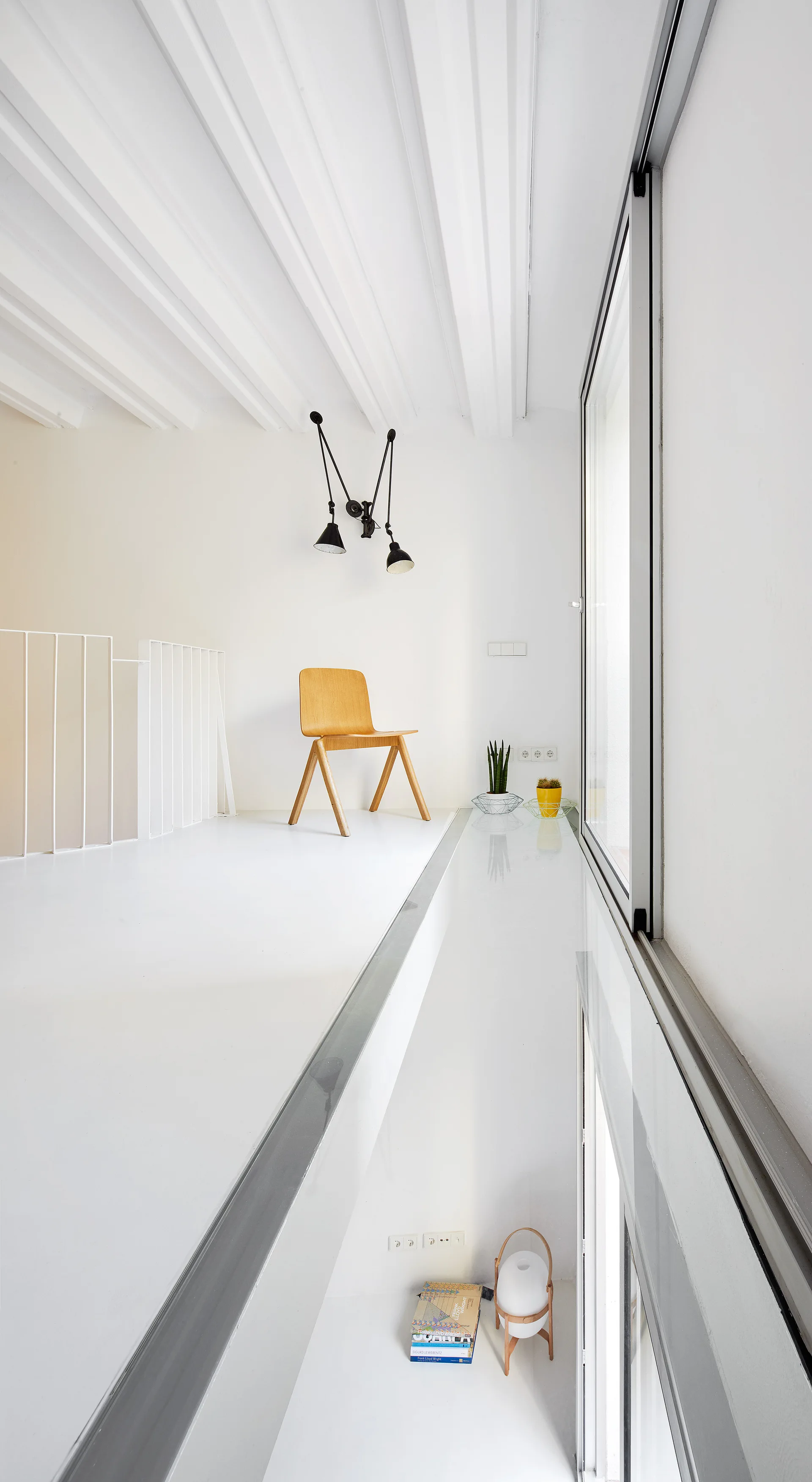A minimalist white mezzanine landing features a natural wood chair, white painted ceiling beams, and an adjustable black lamp.