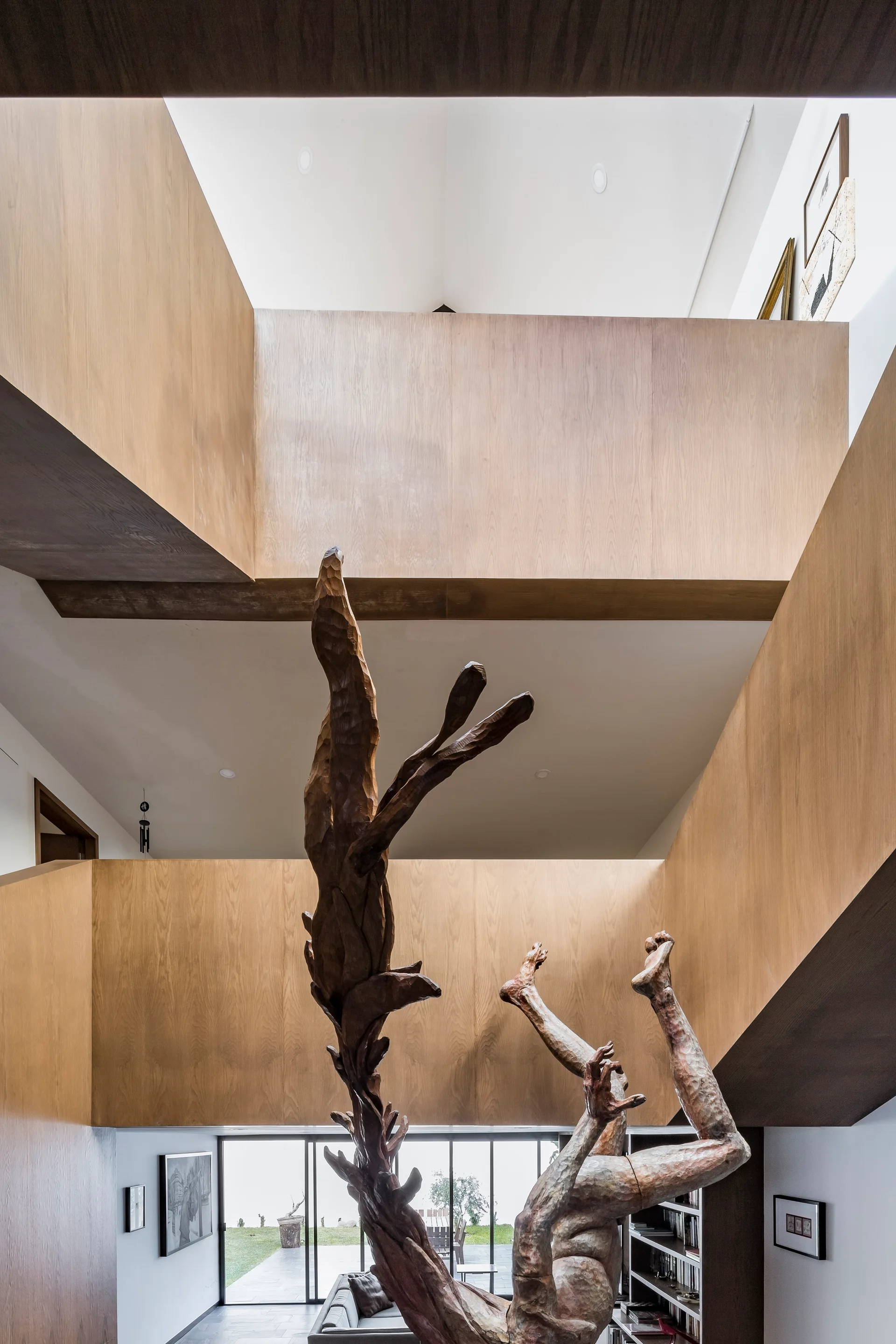 A multi-story atrium features warm wood-paneled walls, an open vertical void, and a tall, abstract wooden sculpture stretching upwards from the ground floor.