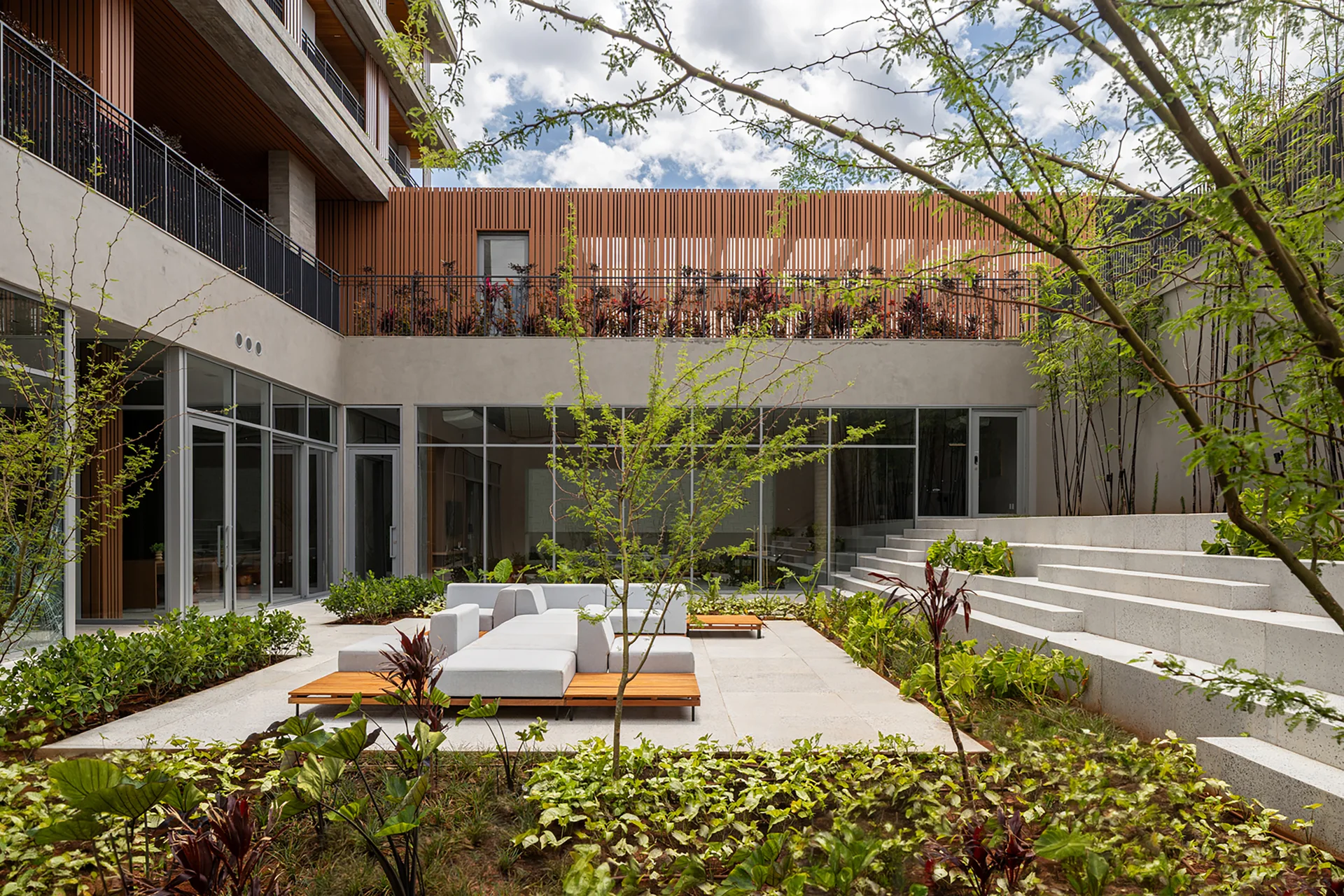 A modern outdoor courtyard featuring a low-profile modular sofa, large stone pavers, tiered concrete steps, and lush green landscaping.