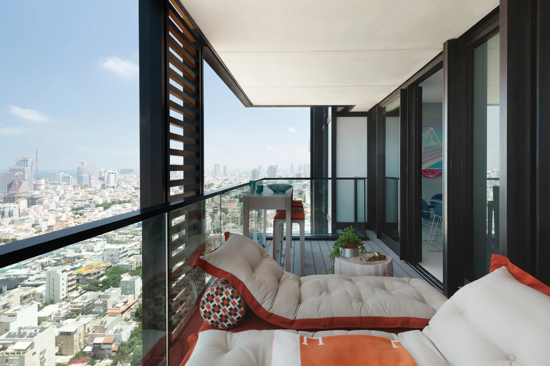 A modern high-rise balcony featuring tufted cream and orange lounge cushions, a glass railing, and a view of the city skyline.