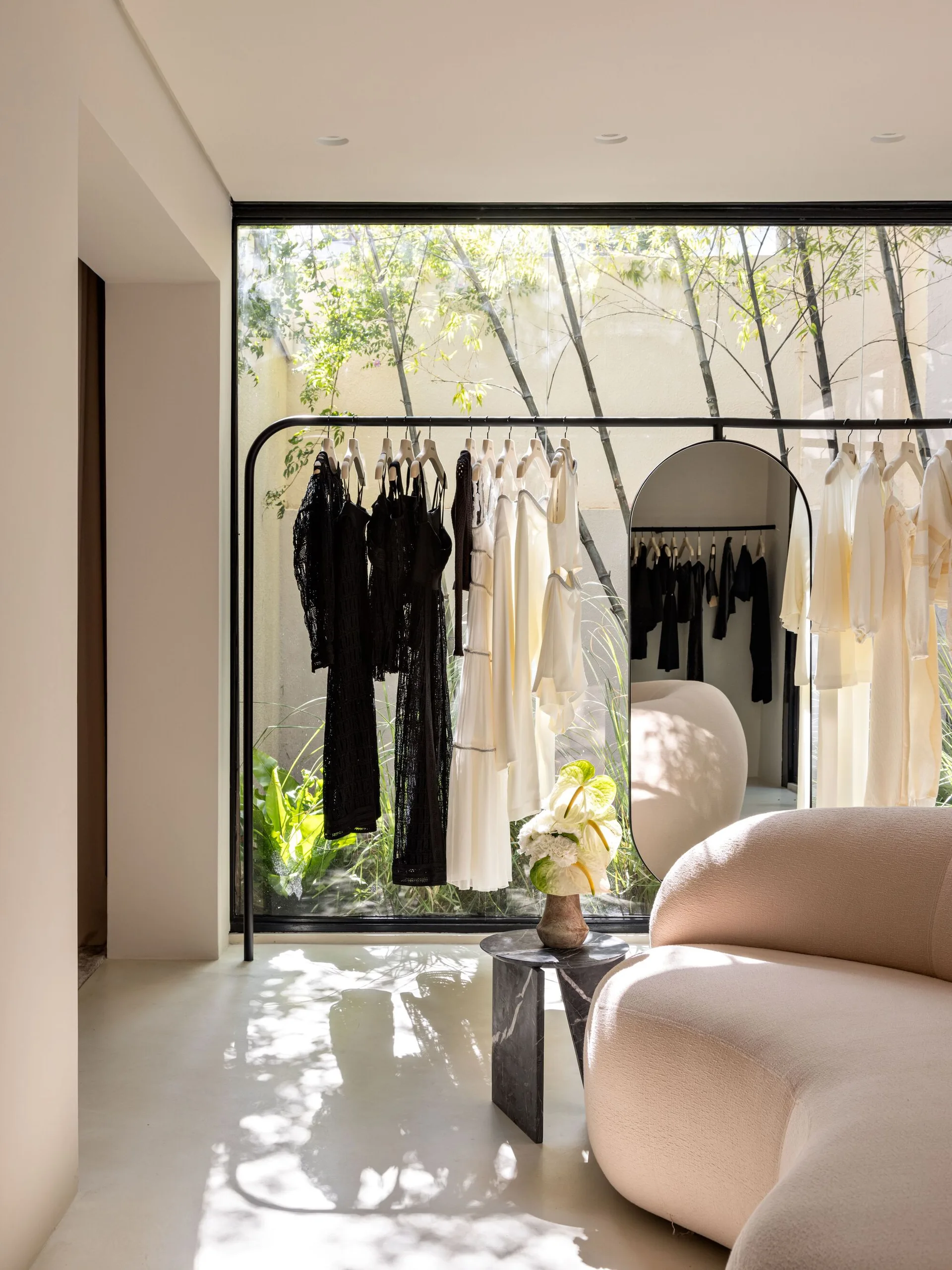 A black clothing rack holding garments stands before a large window overlooking bamboo, next to a curved cream sofa and an arched mirror.