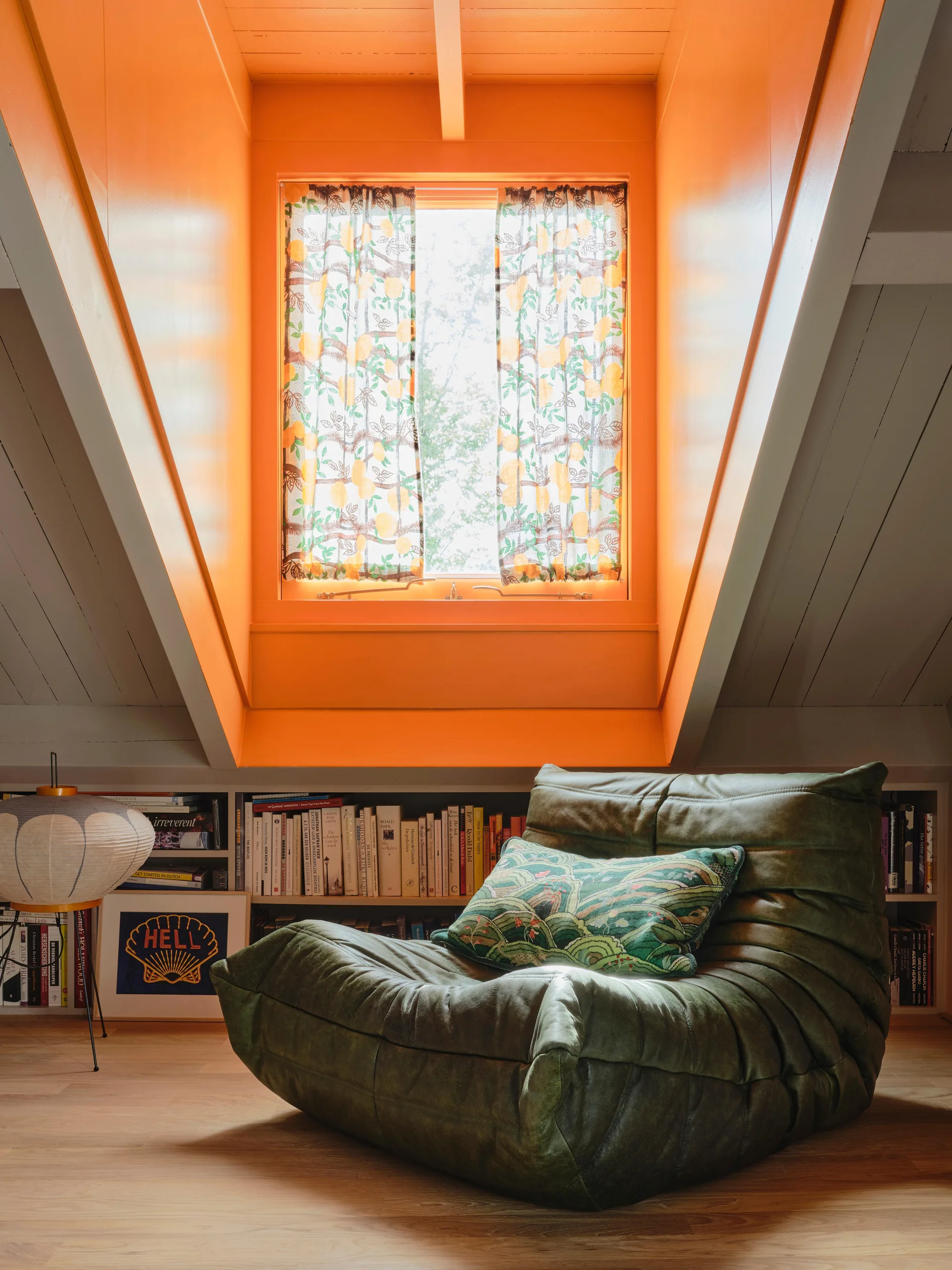 A low green tufted armchair sits on a light wood floor in an attic room beneath a bright orange dormer window and built-in bookshelves.