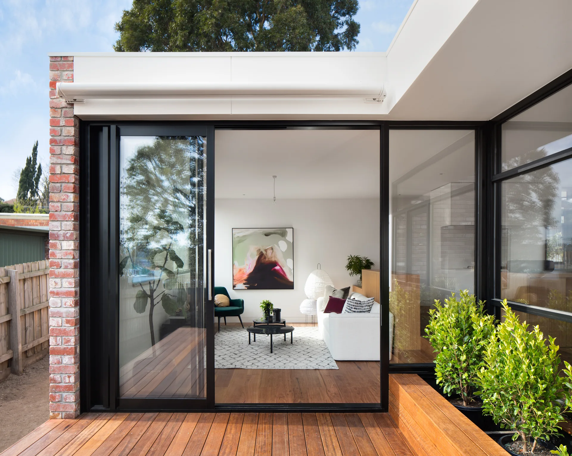 A modern white living room with a sofa and artwork viewed through black sliding glass doors from an outdoor wooden deck.