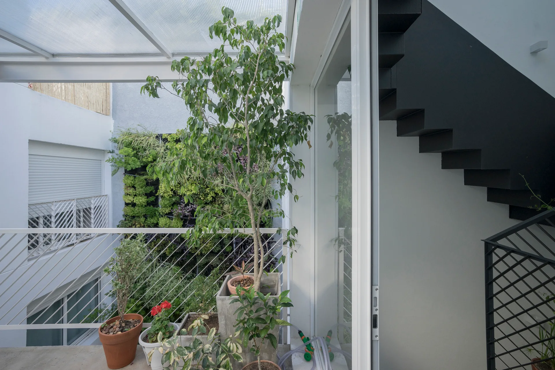 An outdoor terrace features potted plants, a lush vertical garden, a white railing, and a black floating metal staircase under a translucent roof.