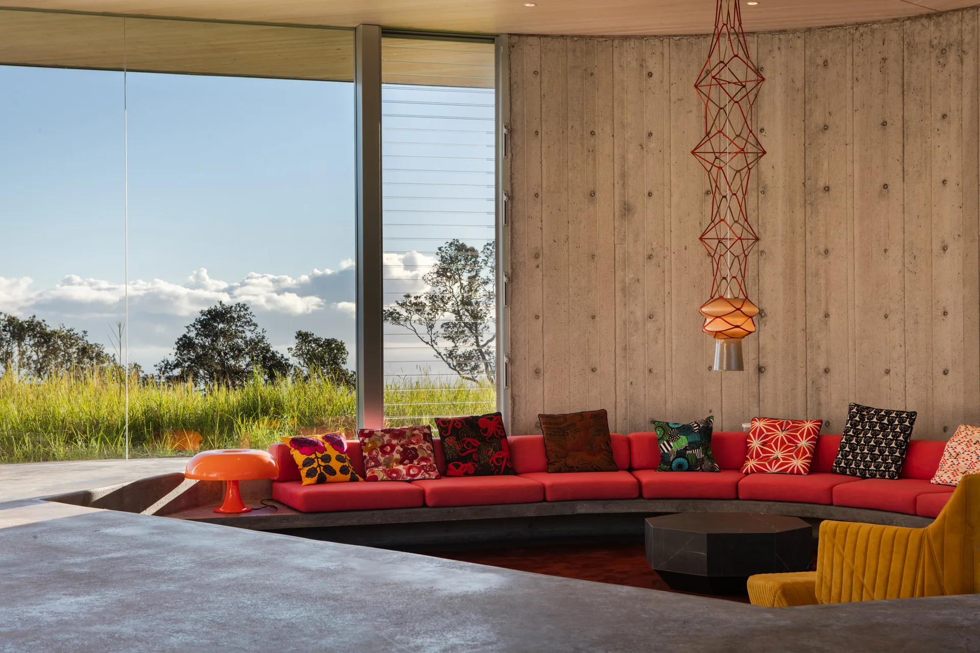 A modern living room features a curved red sofa set against a board-formed concrete wall next to floor-to-ceiling windows.