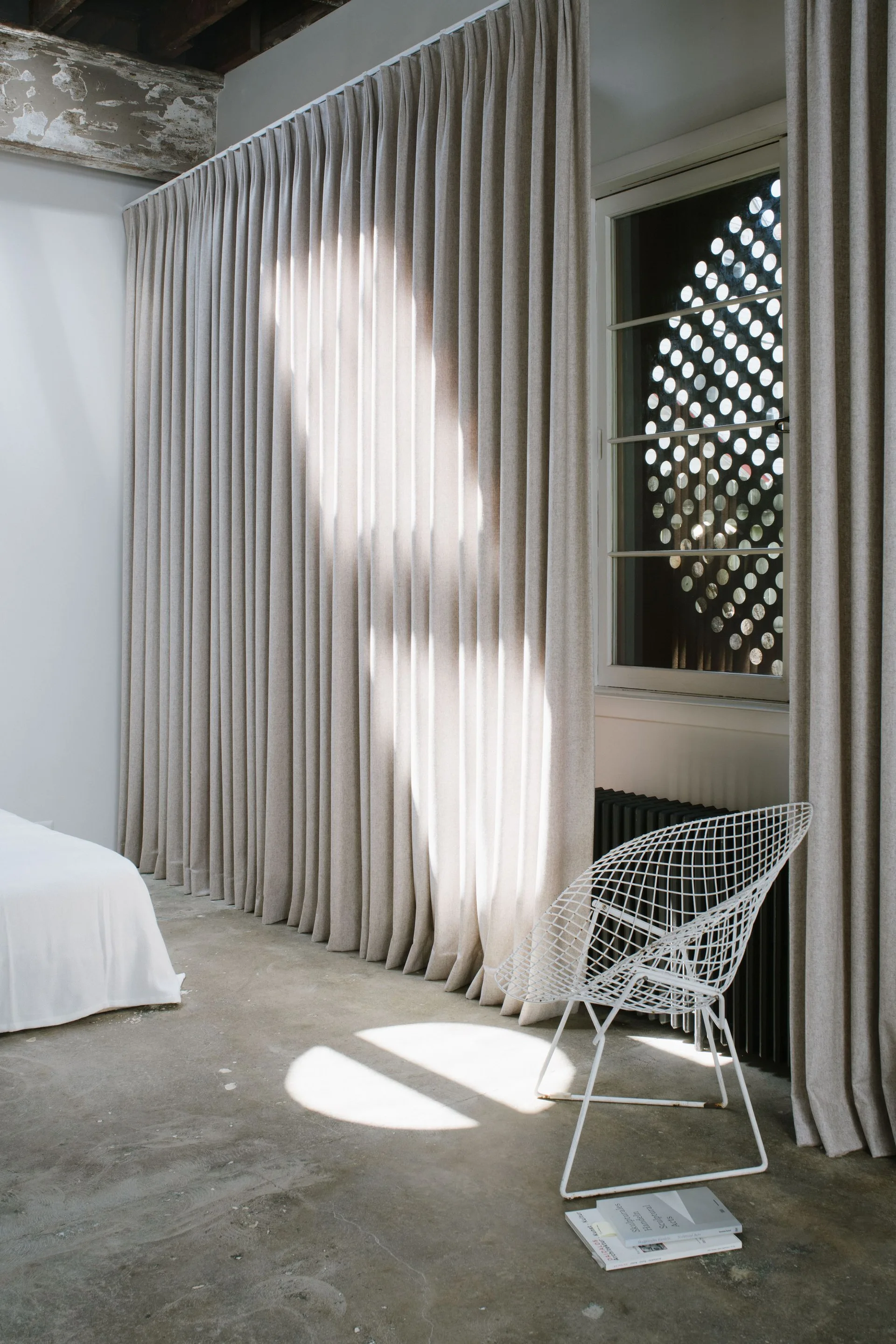 A minimalist bedroom features beige floor-to-ceiling curtains beside a white wire chair on a concrete floor under a patterned window.