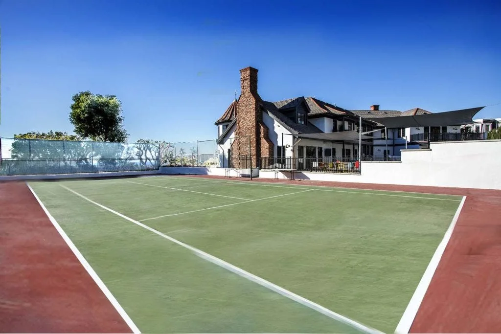 A green and terracotta backyard tennis court sits before a white multi-story house featuring a prominent tall brick chimney.