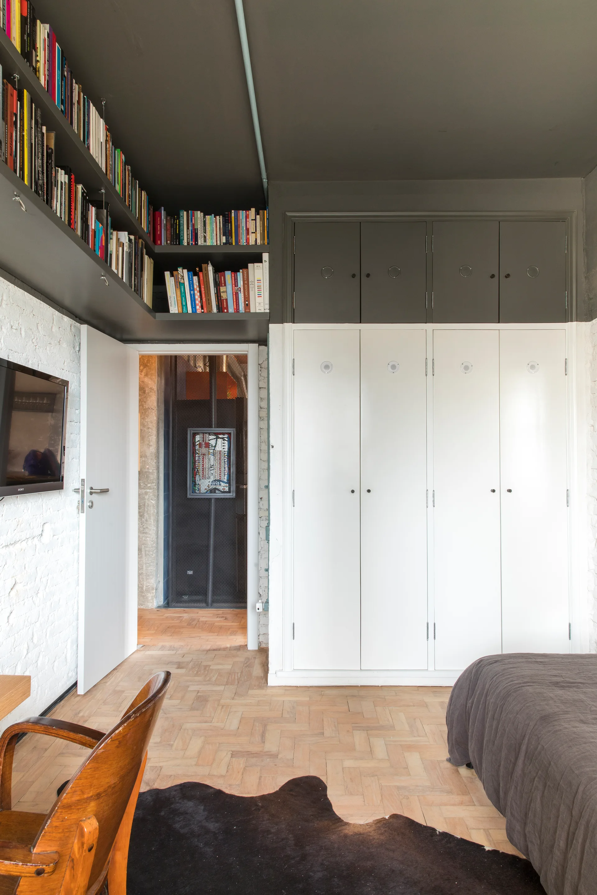 A modern bedroom featuring a dark grey ceiling with built-in bookshelves, white wardrobes, a painted brick wall, and parquet wood flooring.